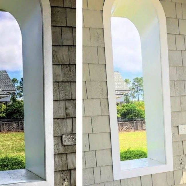 Close-up of a building's exterior wall with two arched openings, showing a backyard with green grass, trees, and neighboring houses in the background.