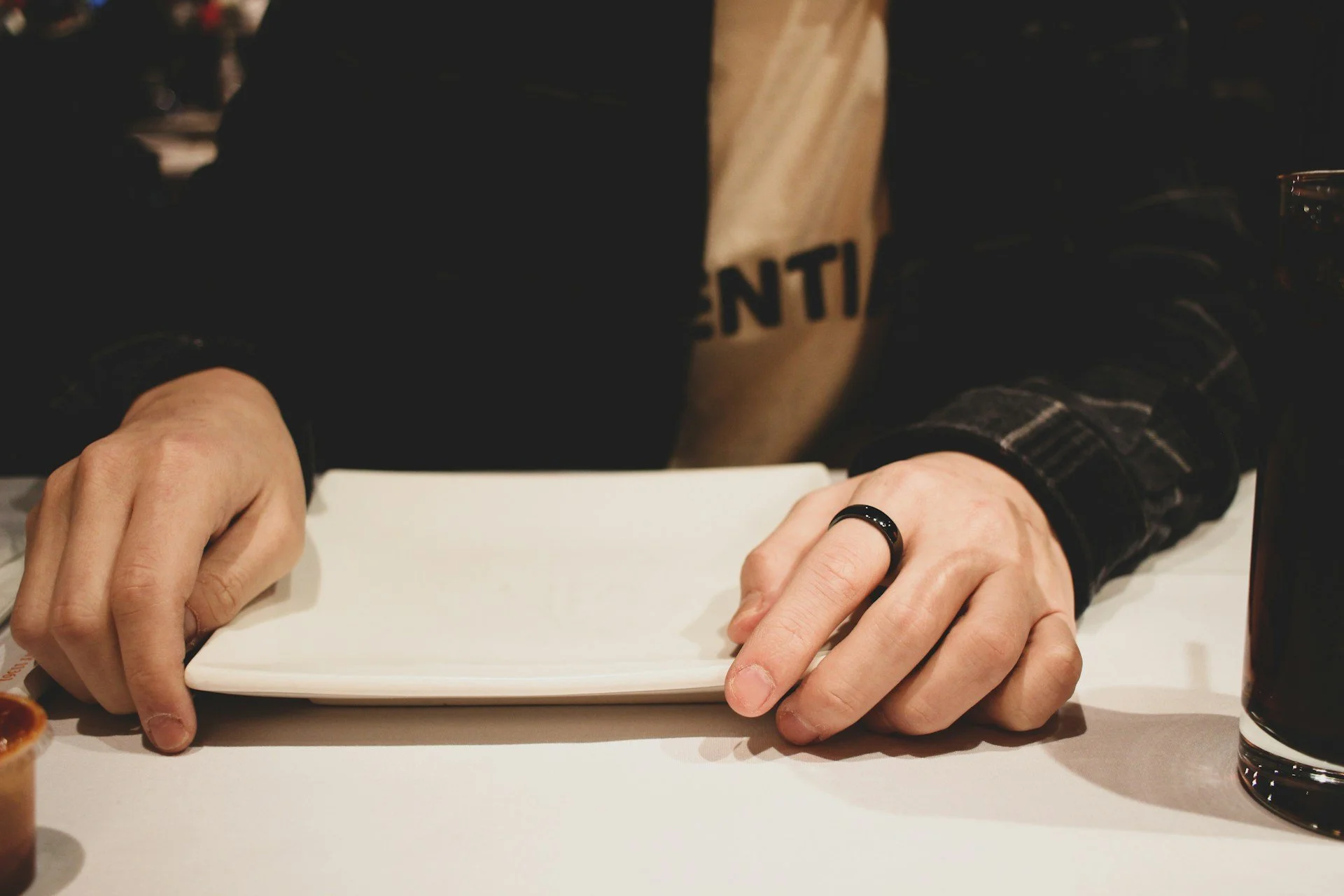 Male torso  in dark jacket and pale yellow t-shirt with black slogan. Hands on table either side of a white square dinner plate. He has a black ring on forefinger of left hand