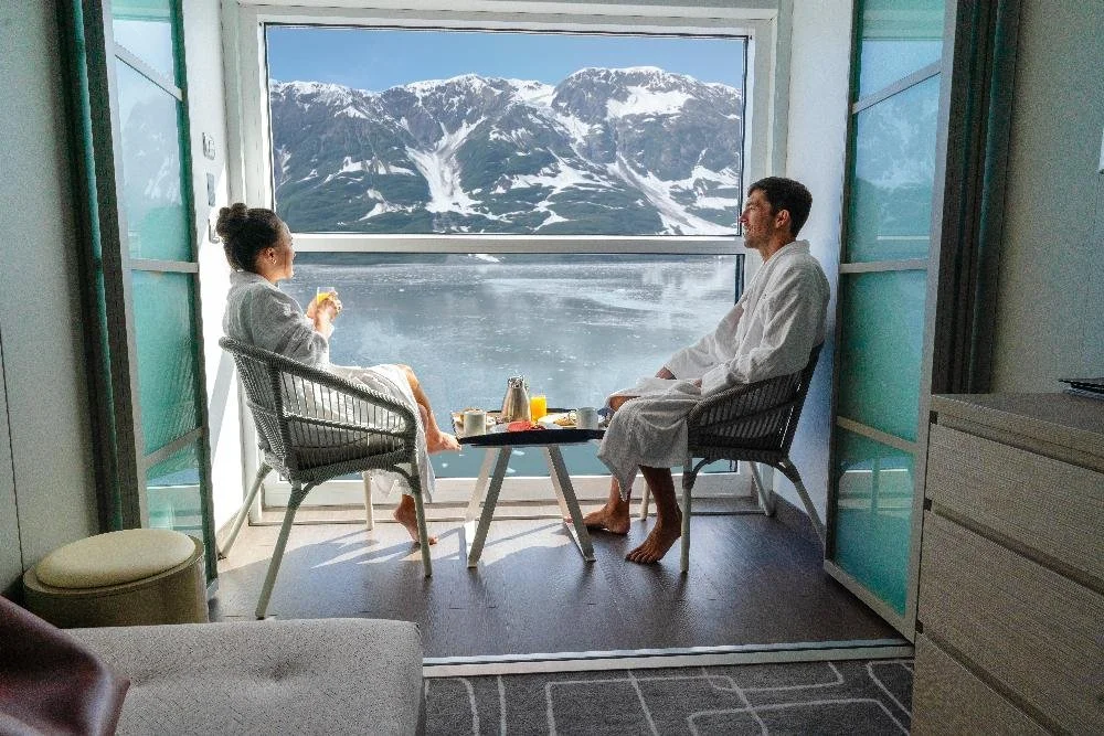A couple enjoying breakfast sitting at their closed infinite veranda on Celebrity Beyond overlooking wintery Alaskan mountains.