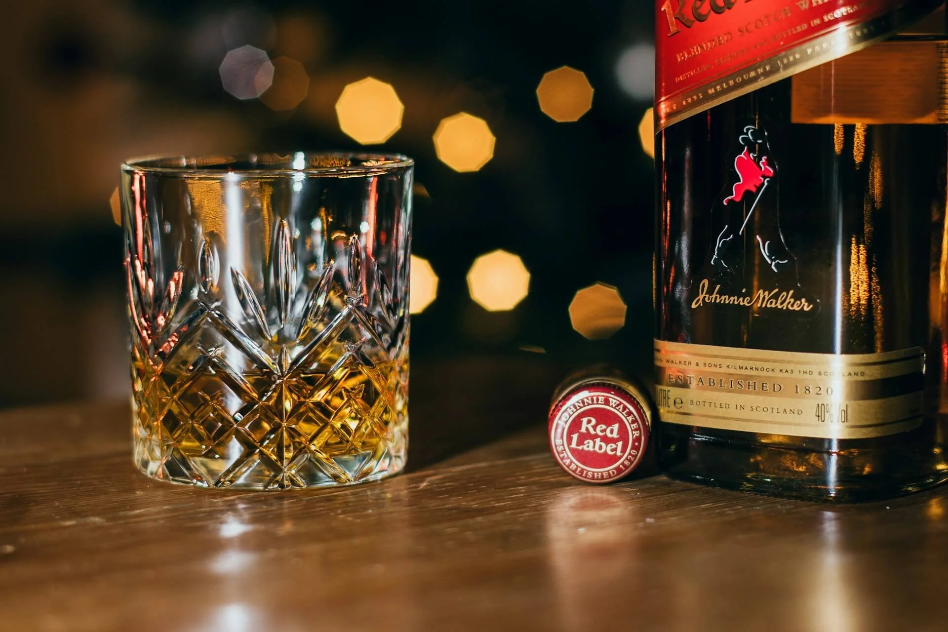 Whiskey tumbler with golden liquid standing on a wooden bar surface alongside a bottle of Johnnie Walker Red Lable whiskey