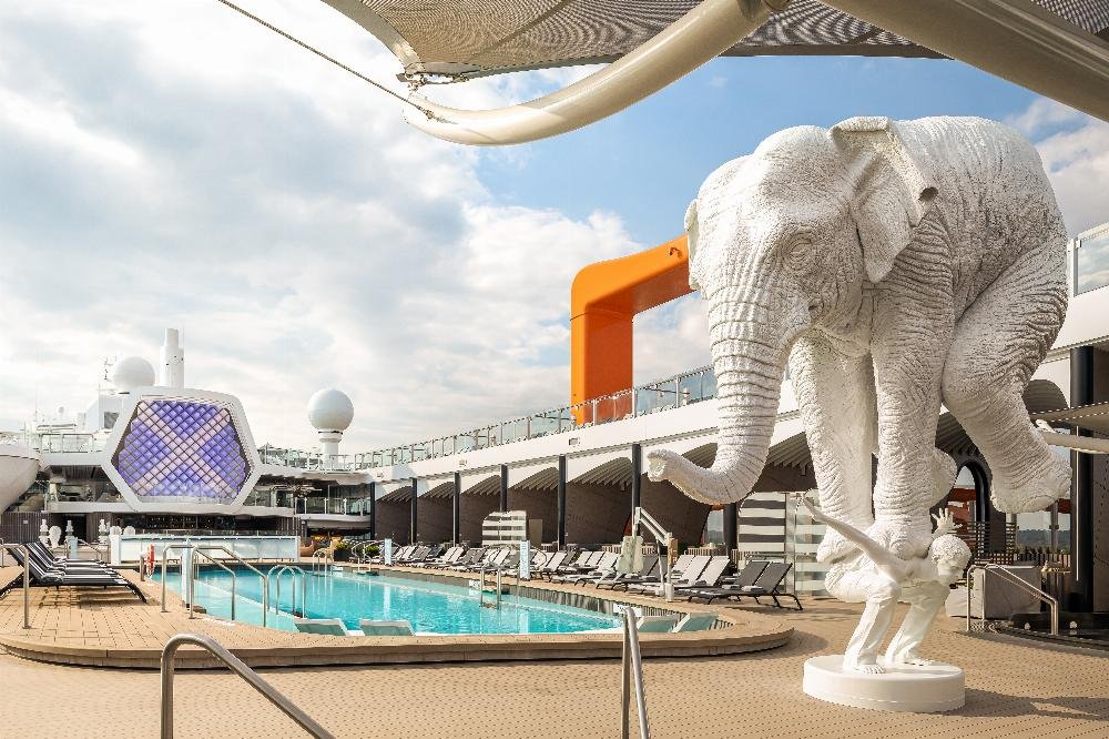 Overview of the Celebrity Beyond Deck 14 resort layout, showing the asymmetric lap pool, sun loungers, the iconic orange Magic Carpet frame, and the signature white elephant sculpture analyzed in our cabin selection guide.