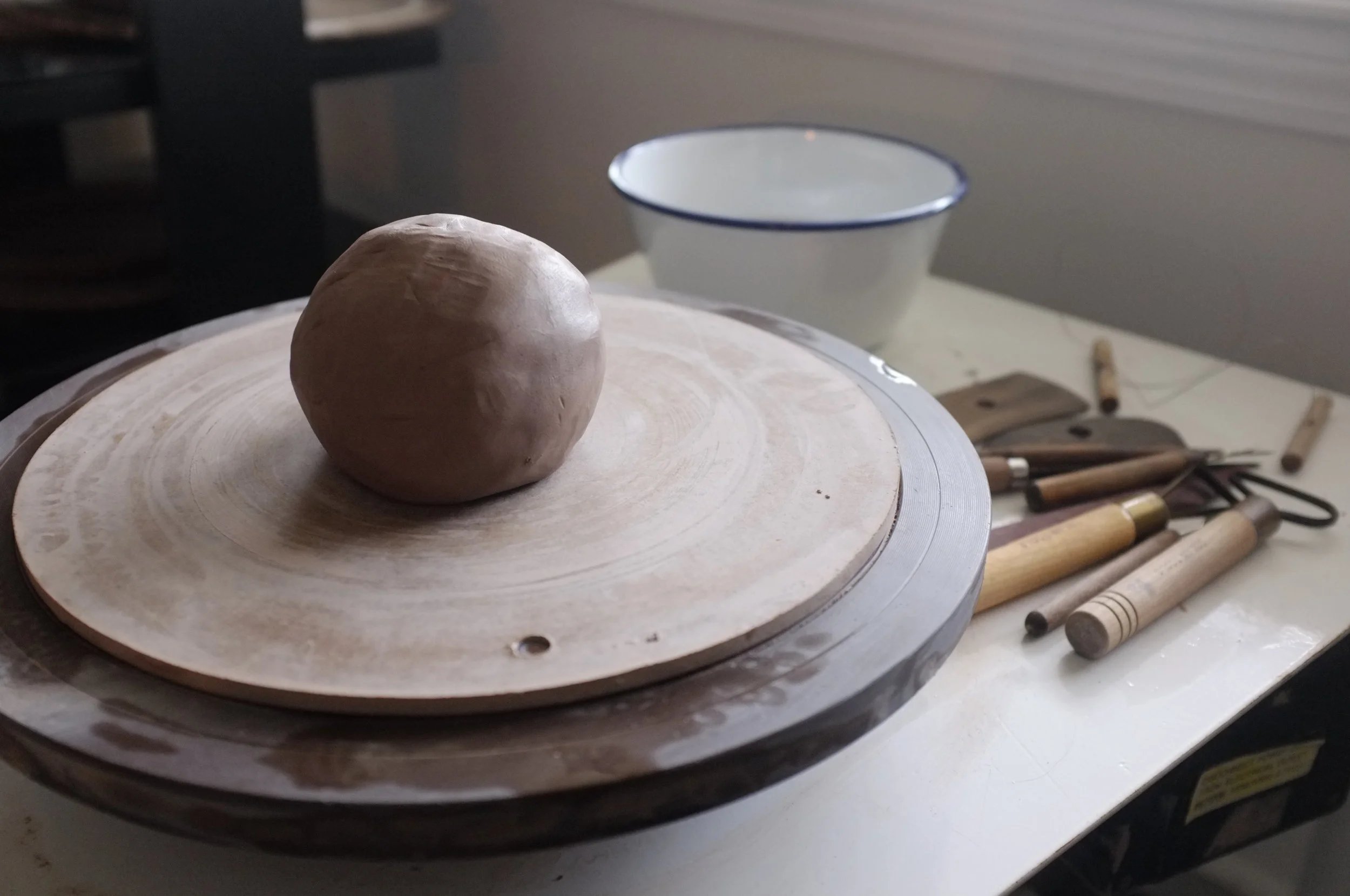 A ball of clay on a pottery wheel, ready to be shaped, with pottery tools and a bowl in the background.