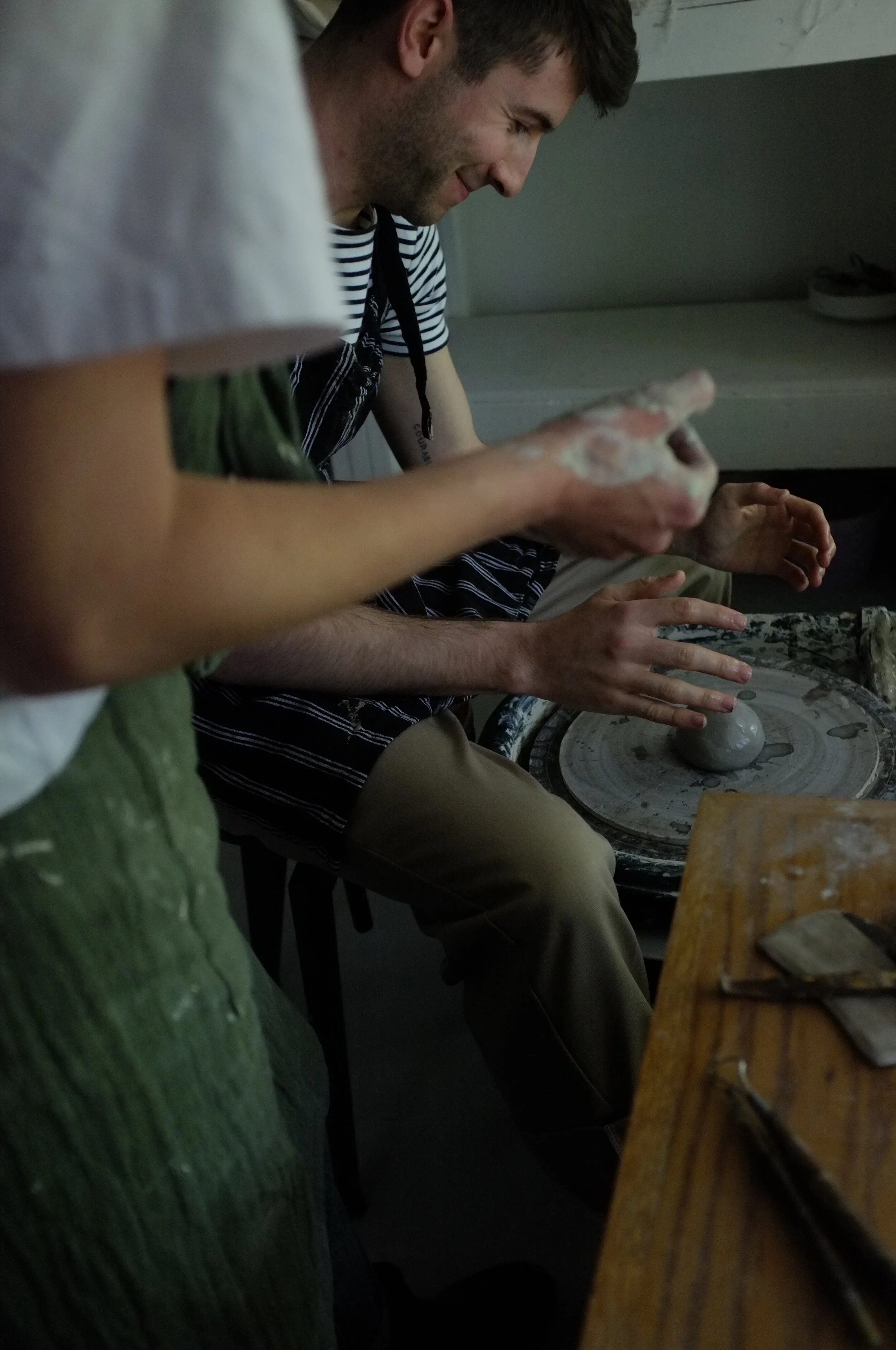 Two people sitting at a table making pottery on a pottery wheel, with one person shaping clay with their hands and the other observing.