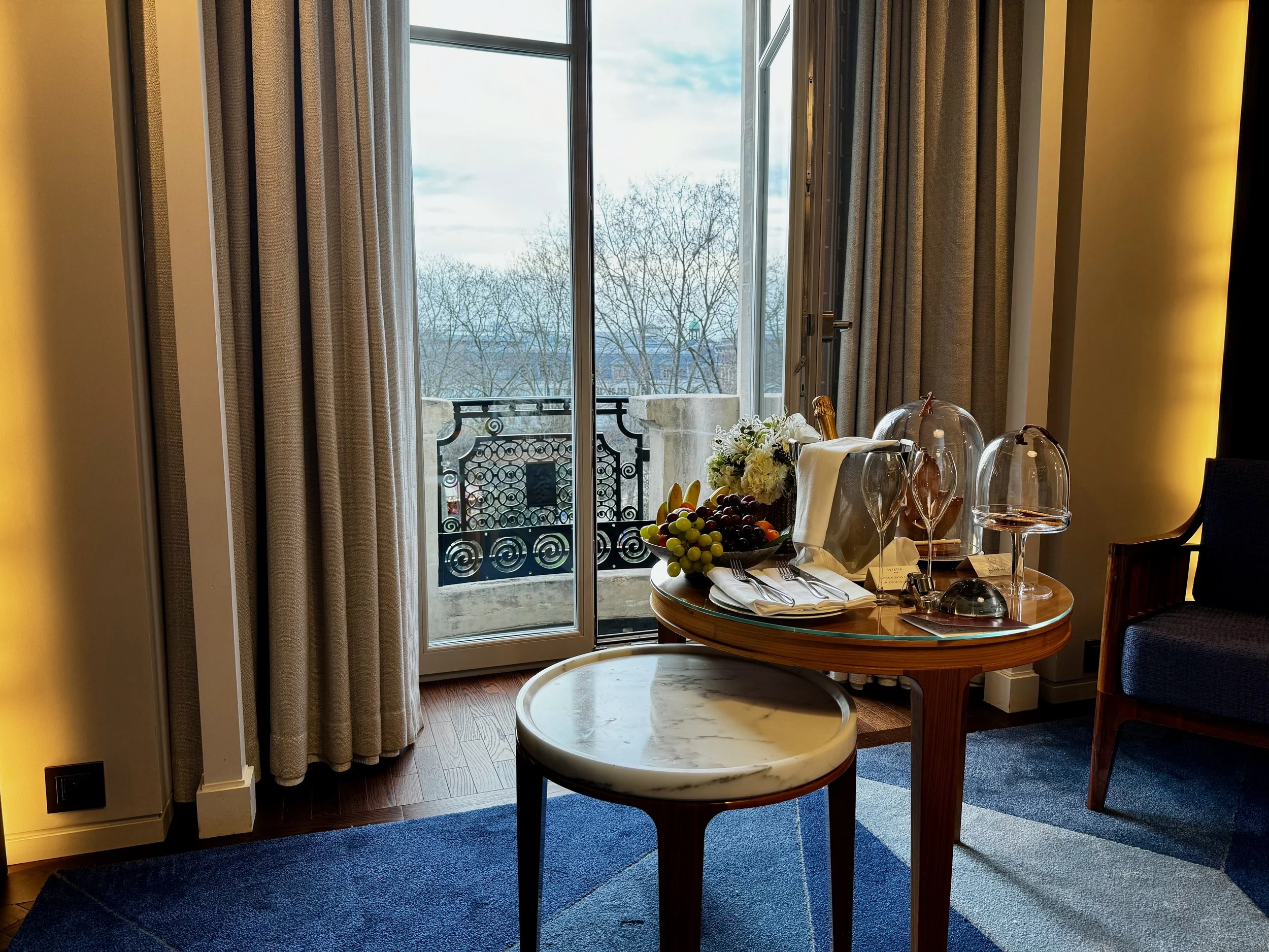 A hotel room with a small round table set with wine glasses, a bottle in an ice bucket, and a floral arrangement, overlooking a balcony with a view of leafless trees outside.