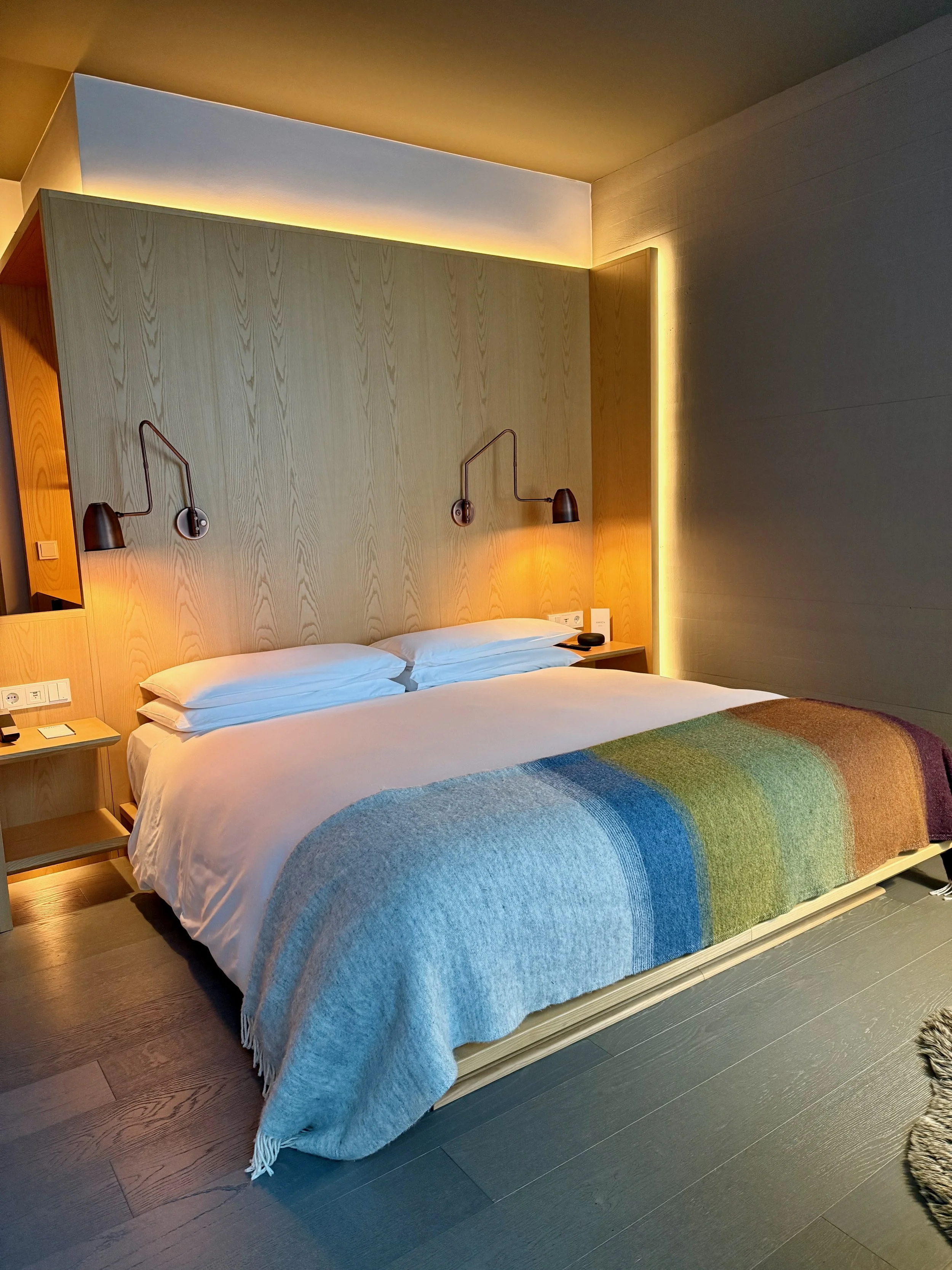 A bedroom with a bed, colorful striped blanket, wooden headboard, and two adjustable wall-mounted reading lamps, with warm lighting.