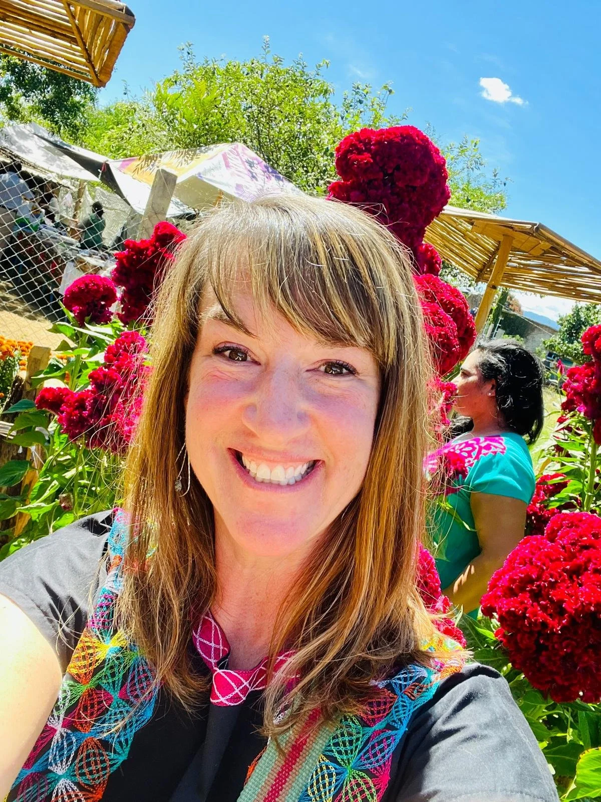 A woman smiling in a garden with bright red flowers under a clear blue sky, wearing a colorful scarf.