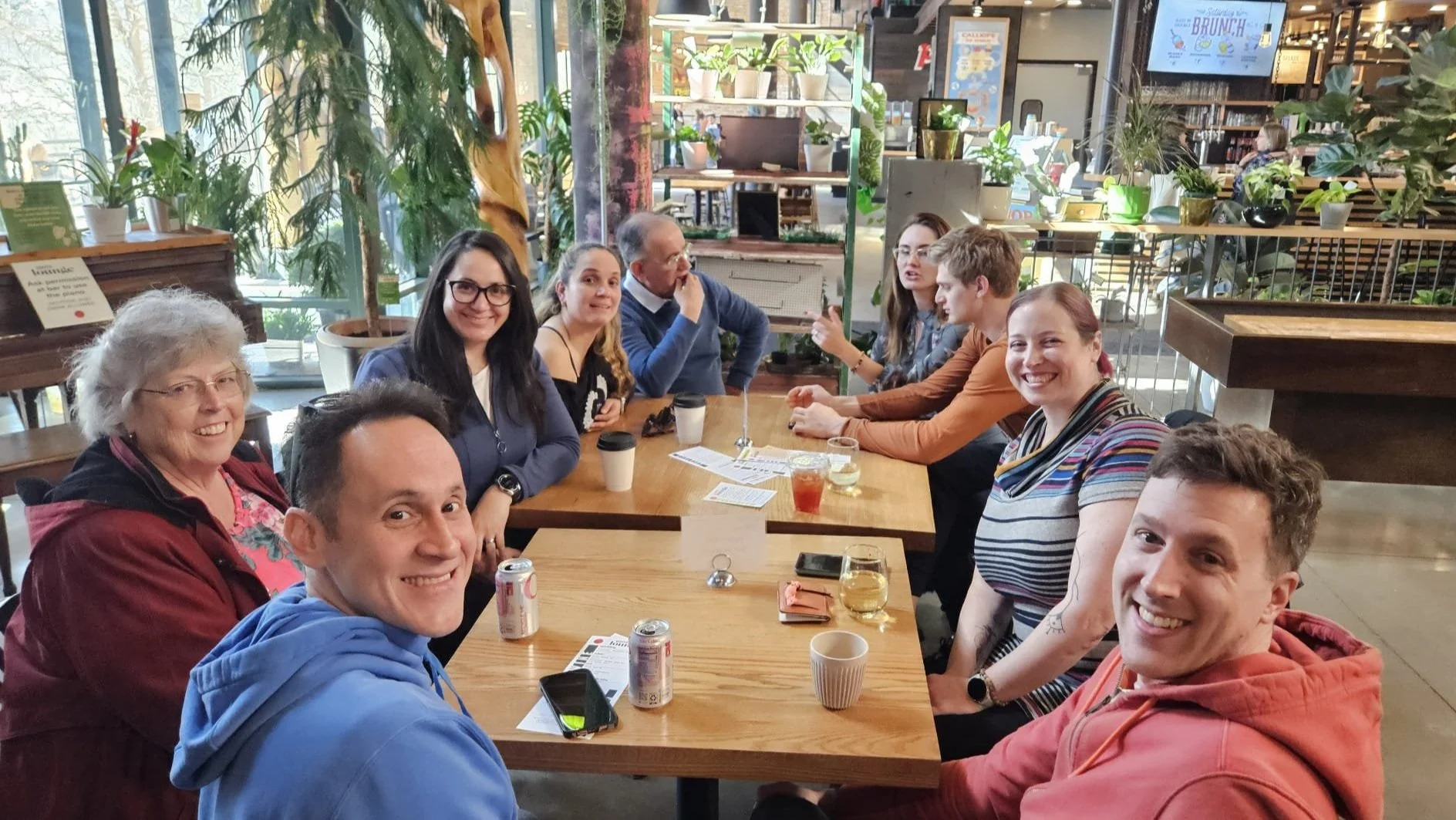 Group of people sitting around a wooden table in a lively, plant-filled cafe, smiling at the camera.