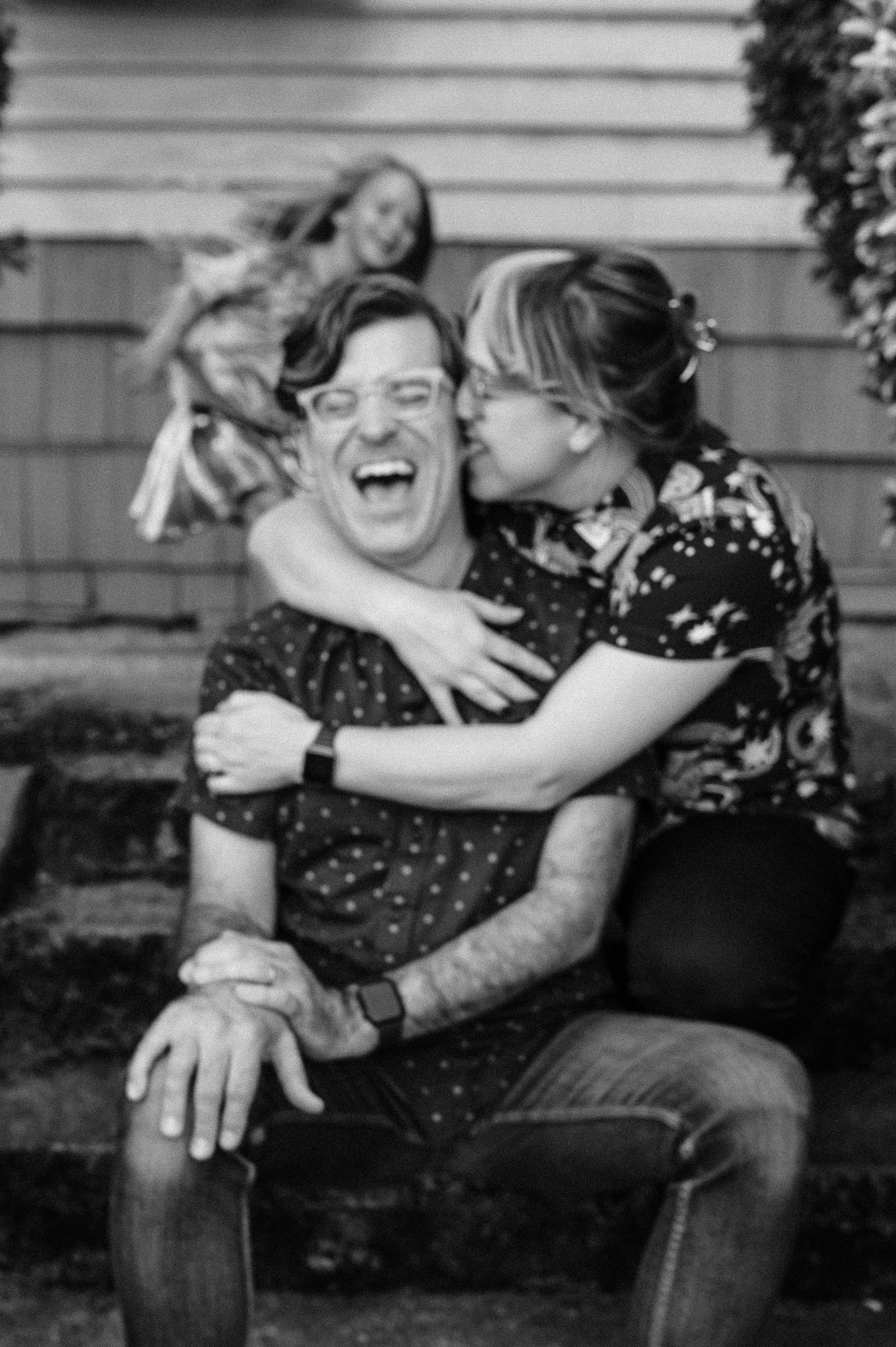 A man wearing glasses and a patterned shirt sitting on a motorcycle, leaning slightly to the right with a woman kissing his cheek. The woman has her hair tied back and is wearing glasses. There is a young girl with her arms around the man, smiling an