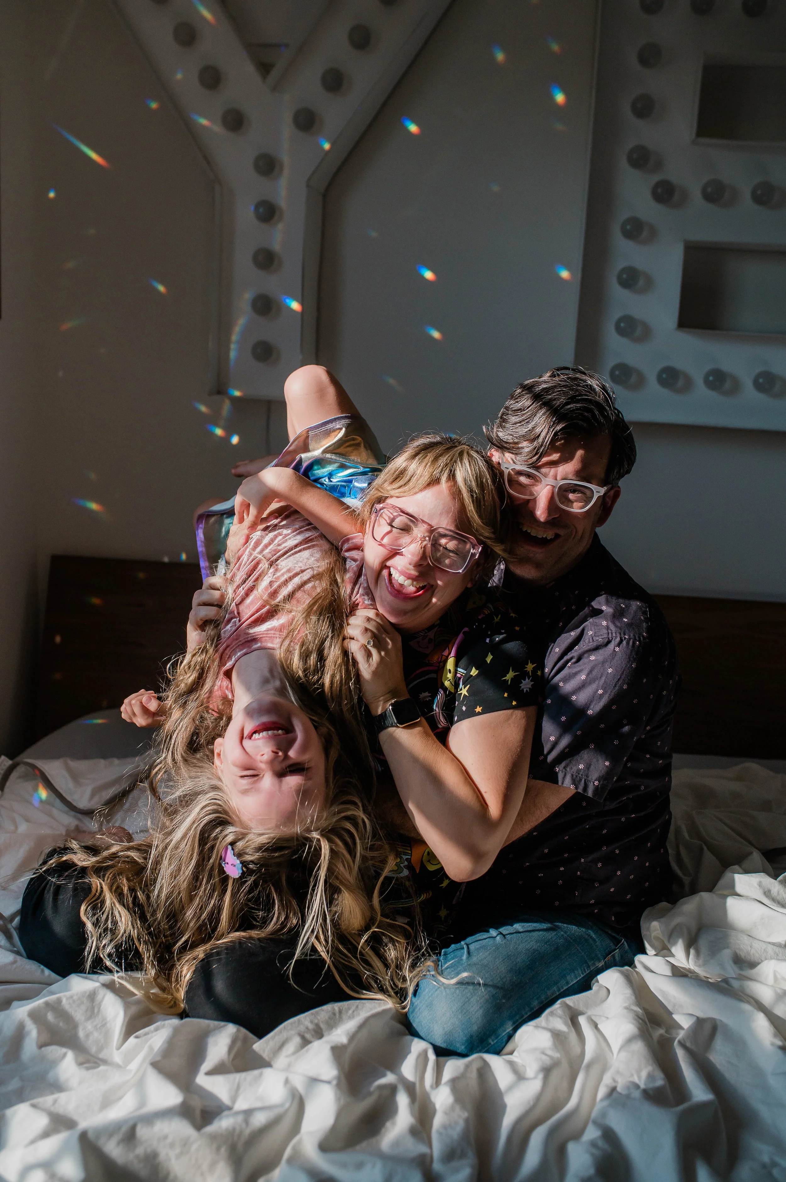 Three people, two women and one man, are laughing and hugging on a bed with crumpled white sheets. A girl with long, curly blonde hair and glasses is lying upside down in the foreground, smiling with her eyes closed. The woman with red hair and glass