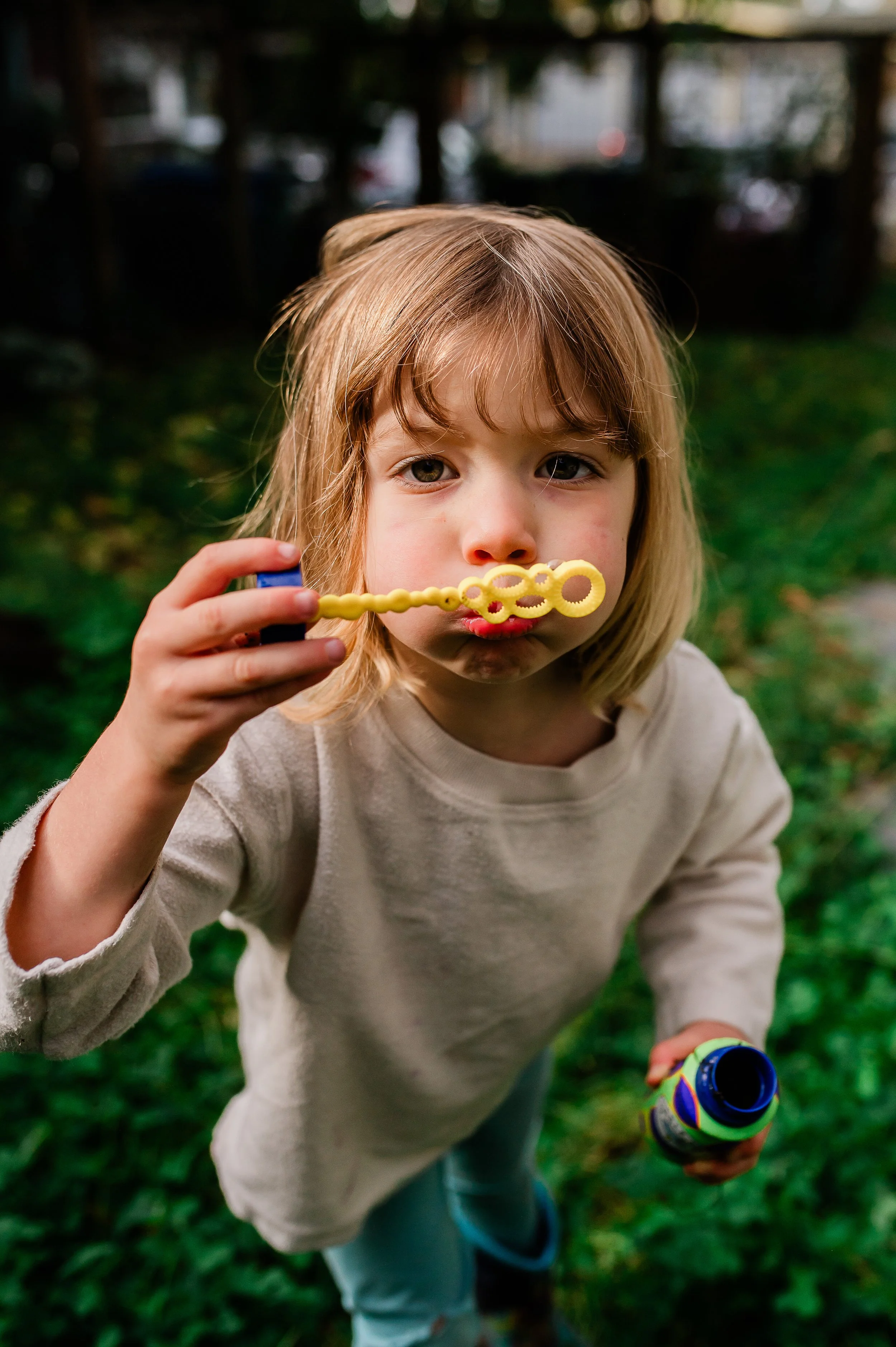 A young girl with blonde hair holds a bubble wand to her lips while standing outside. She appears to be blowing bubbles, with a bubble bottle in her right hand. The background is a yard with grass and trees, and some blurred structures or vehicles.