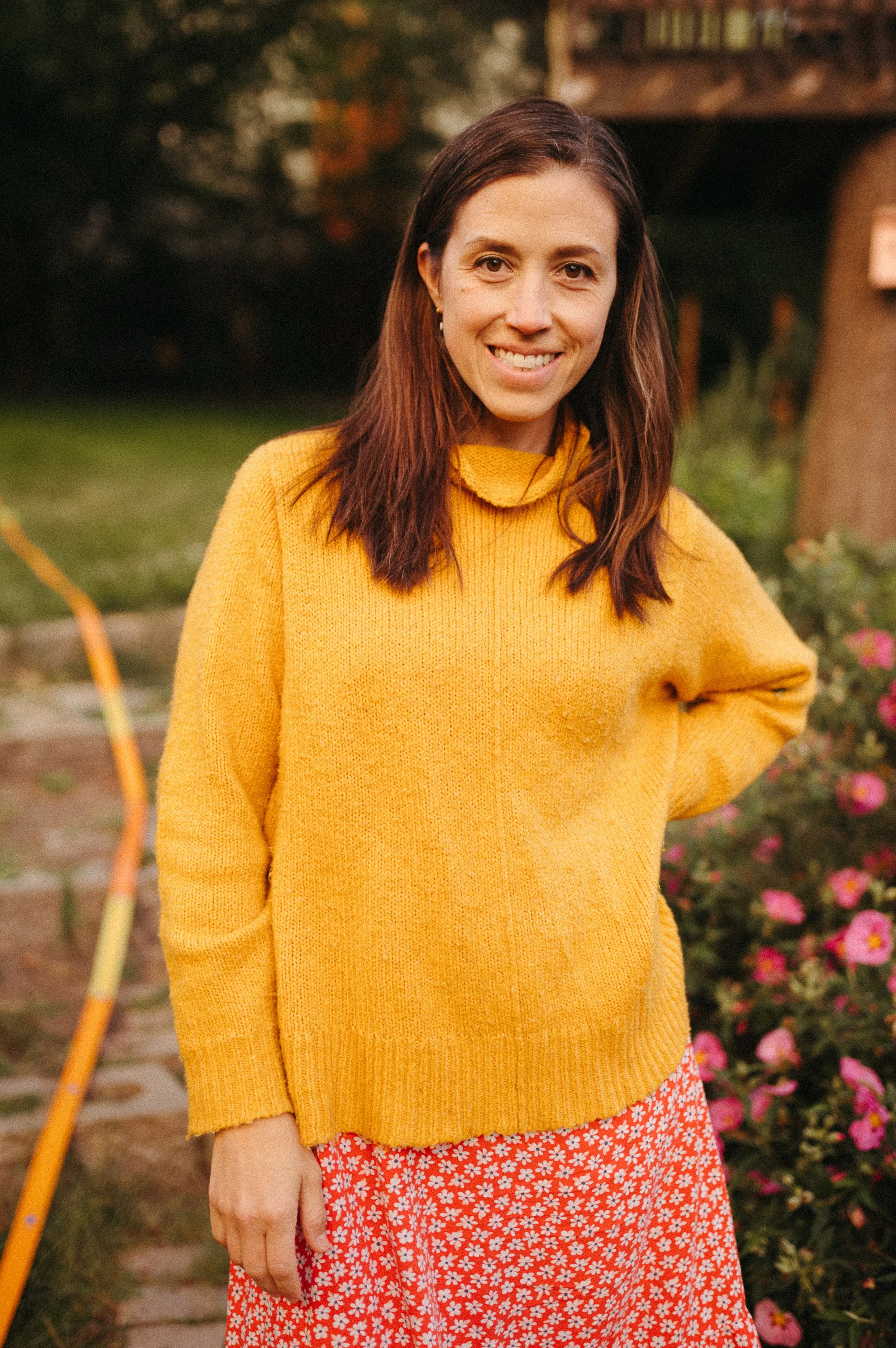 A woman in a yellow sweater and a red floral skirt standing outdoors surrounded by flowers and greenery.