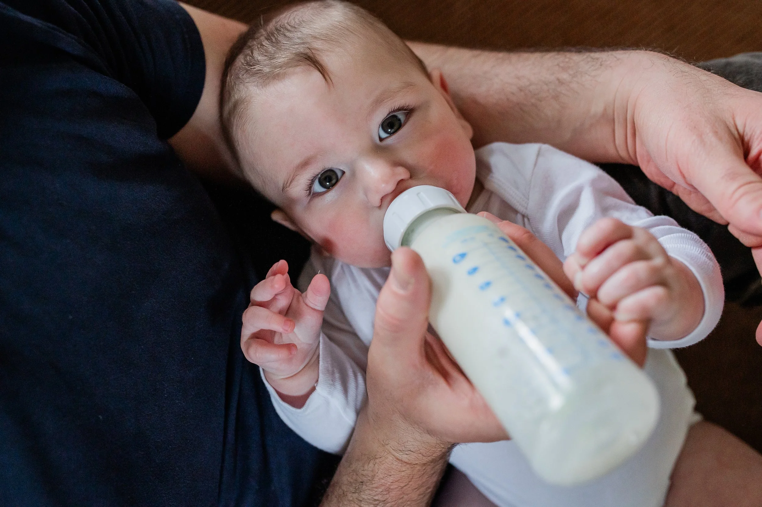 A young child with blue eyes and light hair lying on an adult's lap, drinking from a baby bottle.