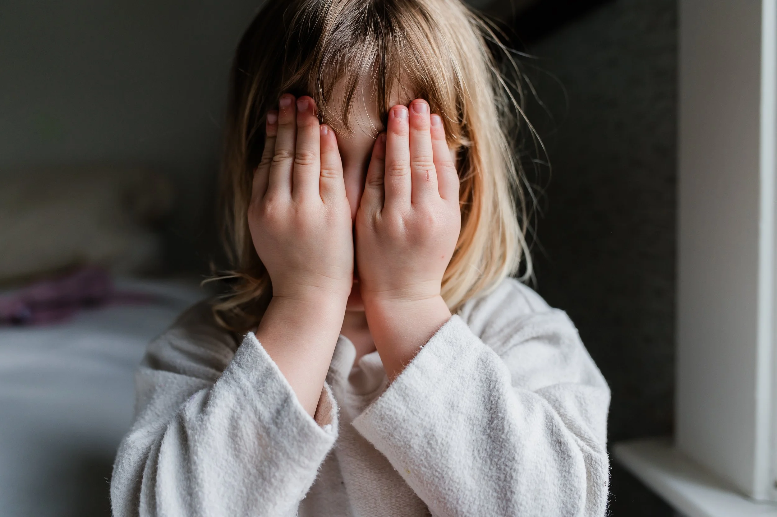 A young girl with blonde hair covering her face with her hands, standing indoors near a dark wall and a light-colored window or door.