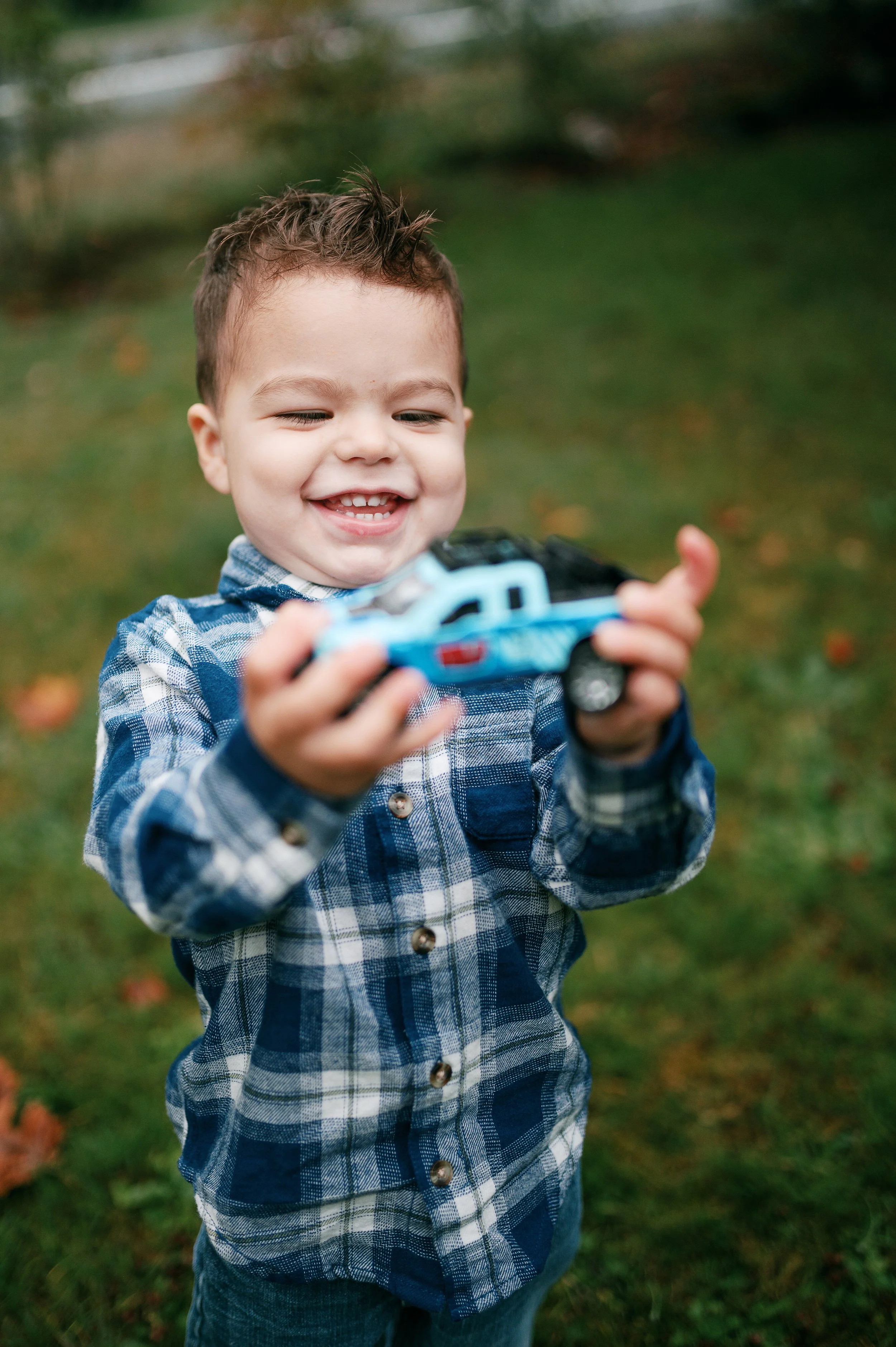 A young boy with short curly hair wearing a blue plaid shirt holding a toy race car outdoors on green grass.