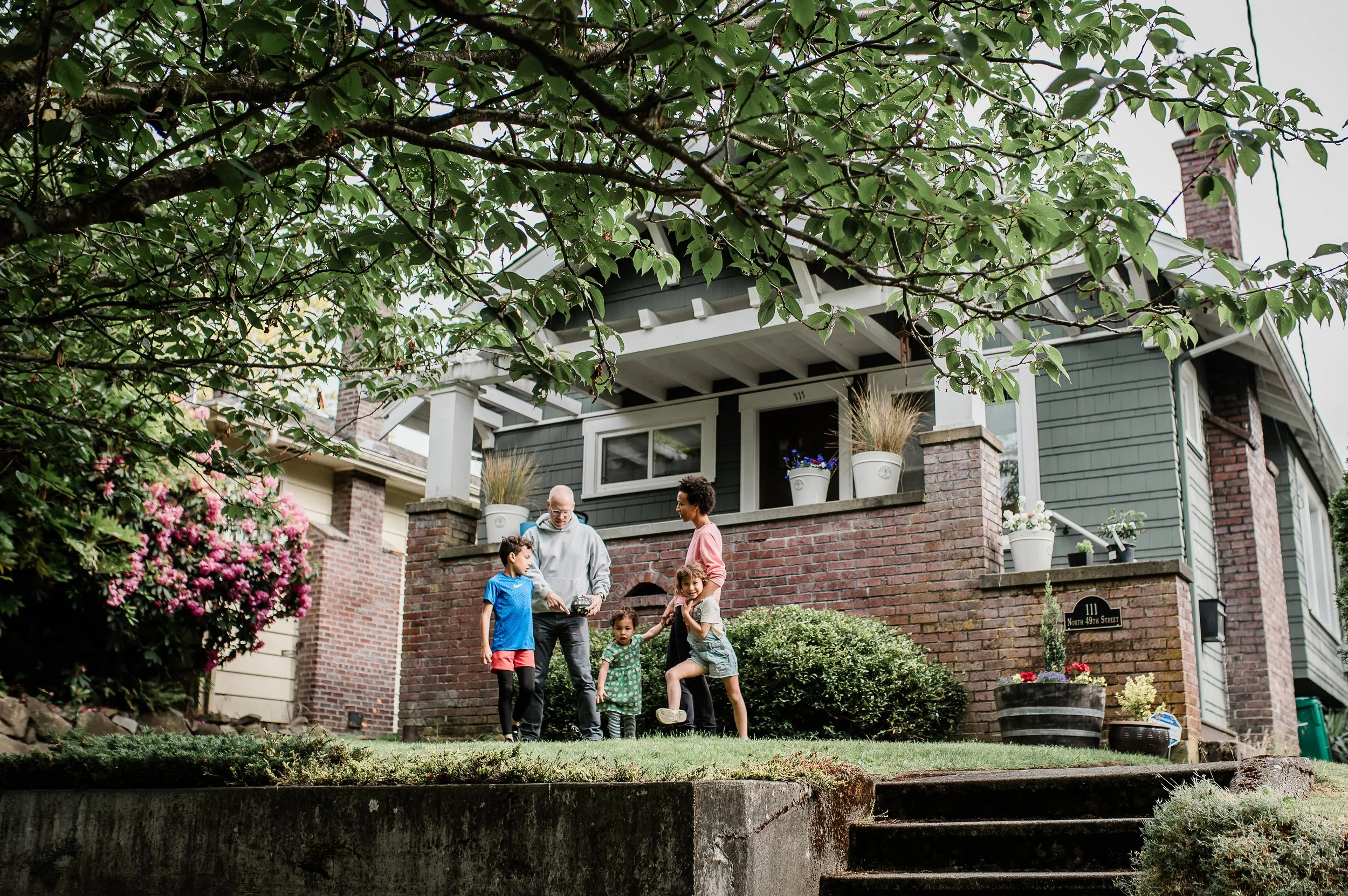 A family with children gathered on the front steps of a house, with lush greenery and flowering plants surrounding them.