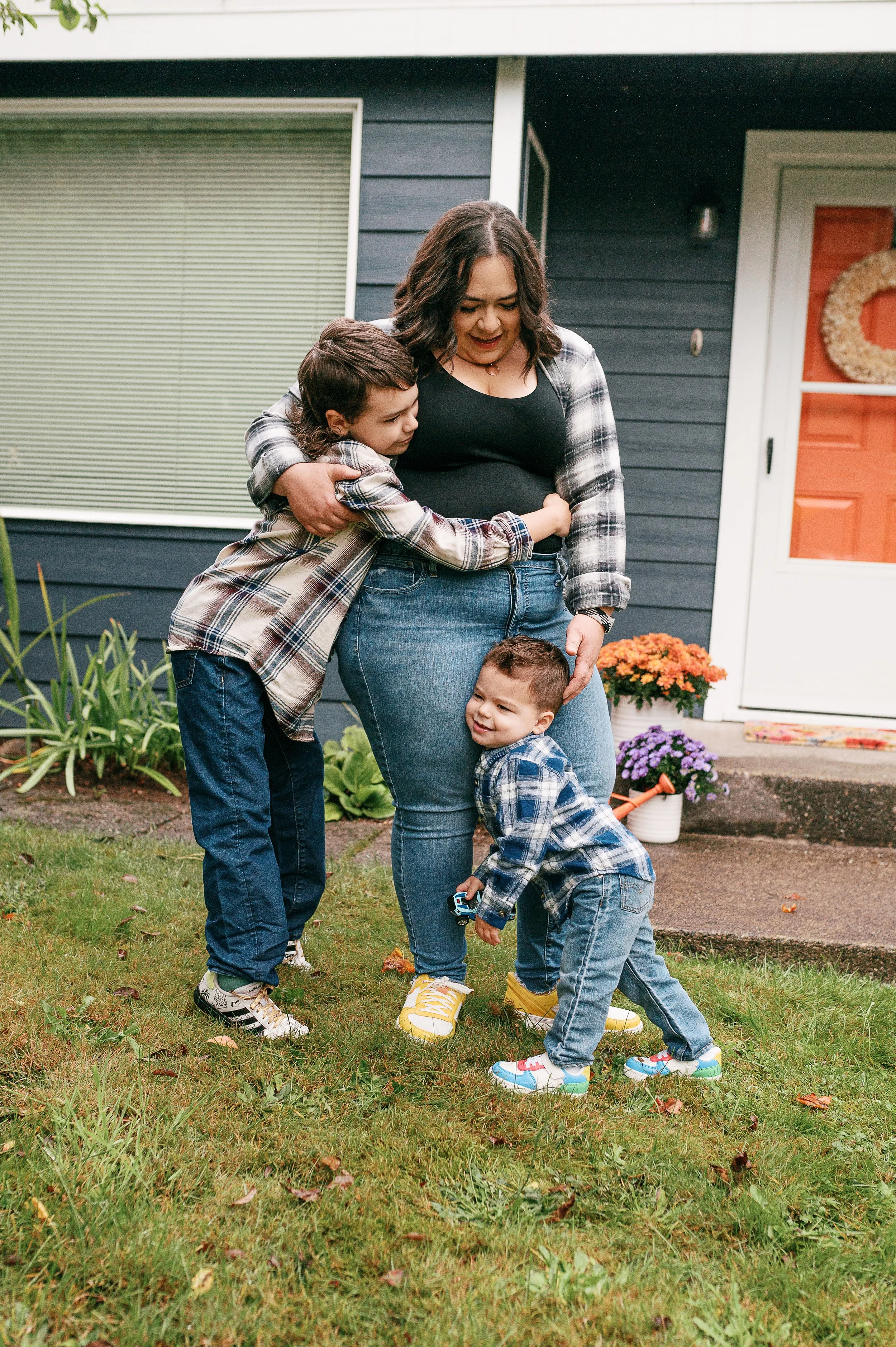 A woman and three young boys hugging and playing outside in a front yard in autumn. The woman is wearing a black shirt, blue jeans, and yellow shoes. The boys are wearing plaid shirts, blue jeans, and colorful sneakers.