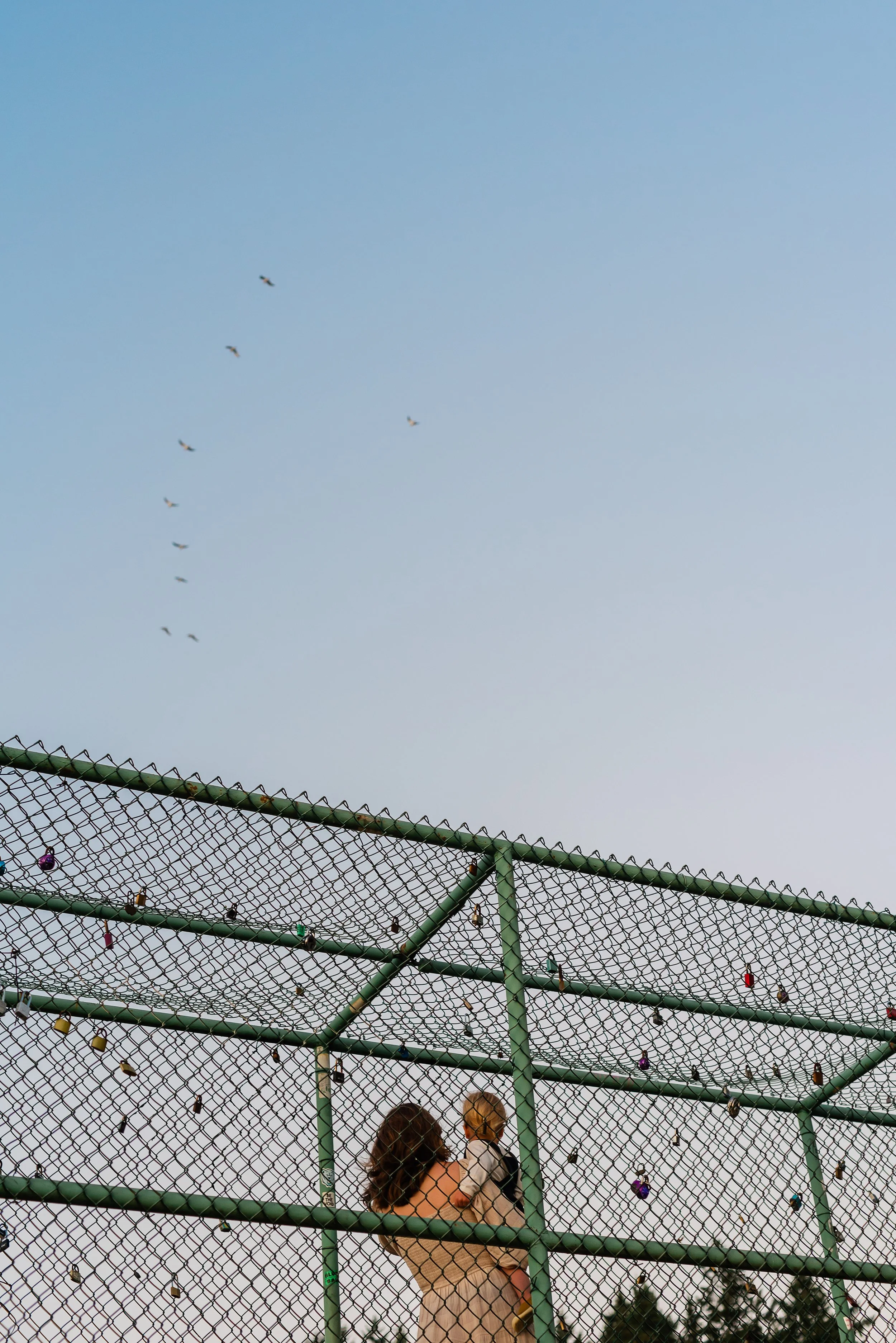 Two women and a child behind a green chain-link fence decorated with colorful locks and ornaments, with a light blue sky and a few birds flying overhead.