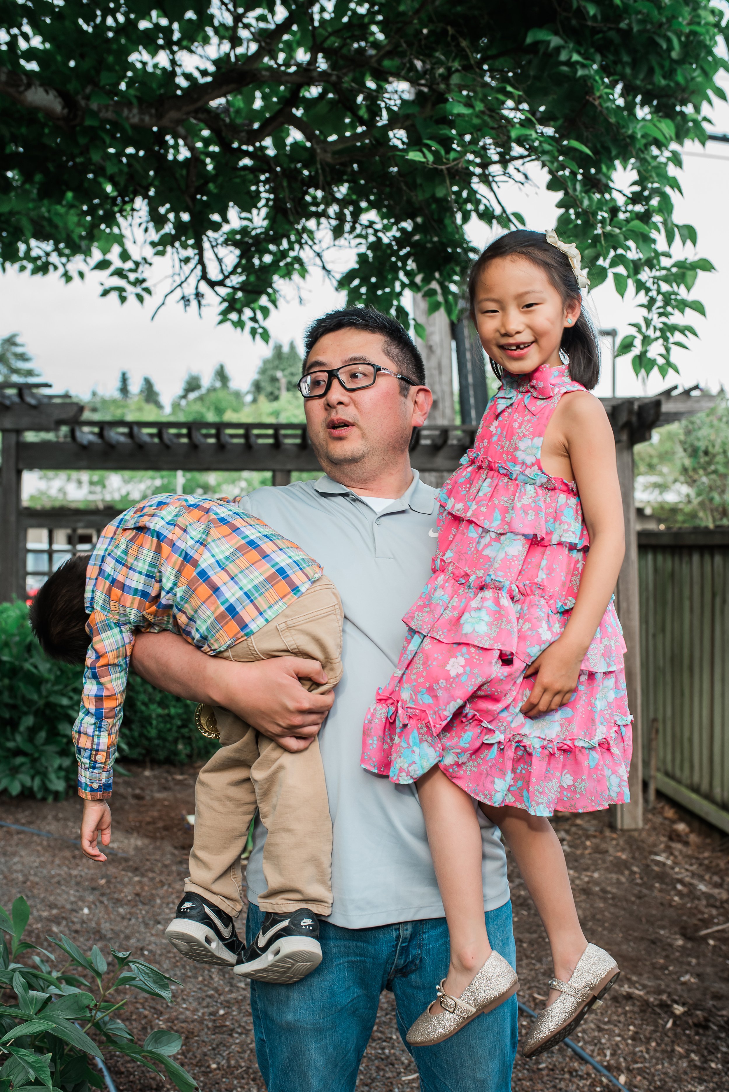 A man holds a young girl on his shoulders while another child is intertwined around his waist. They are outdoors, under a large tree, in a garden or backyard setting.