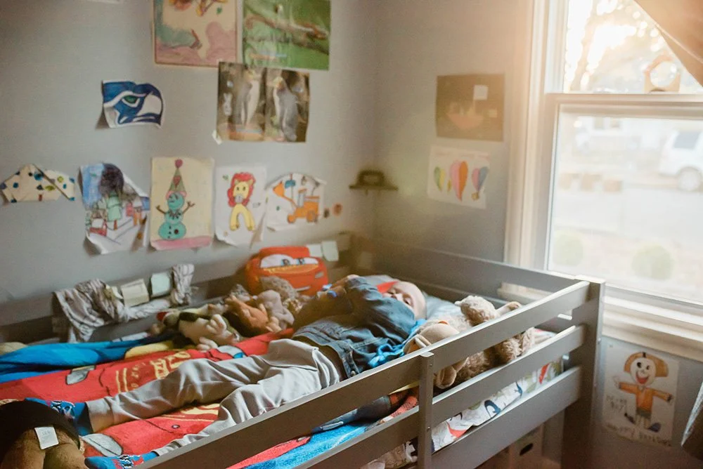 A child lays on his bed in beautiful light with  child made art on the walls.  Anela Deisler Seattle Family Photography