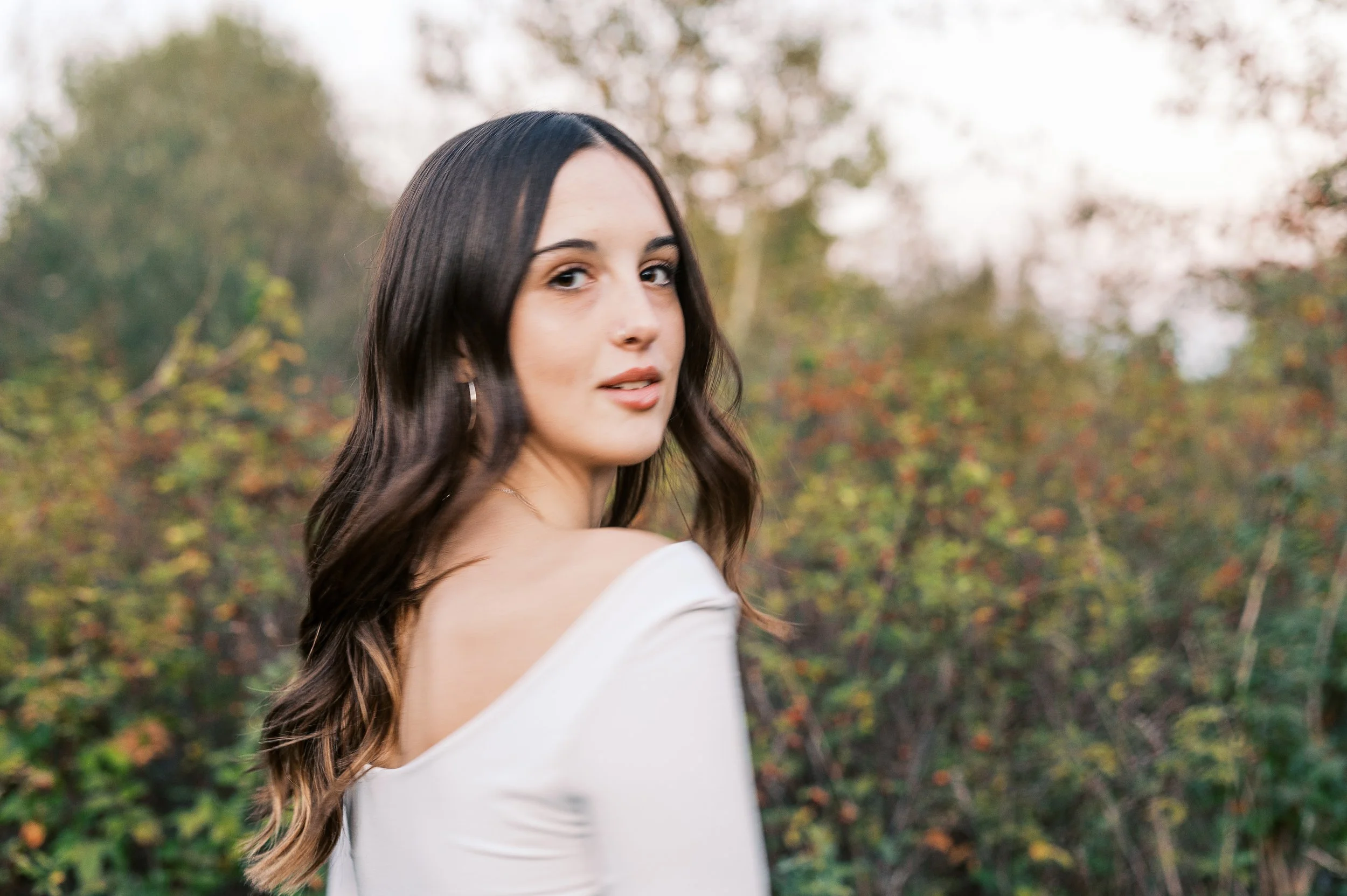 Young woman with long wavy hair looking over her shoulder outdoors with blurred autumn trees in the background.