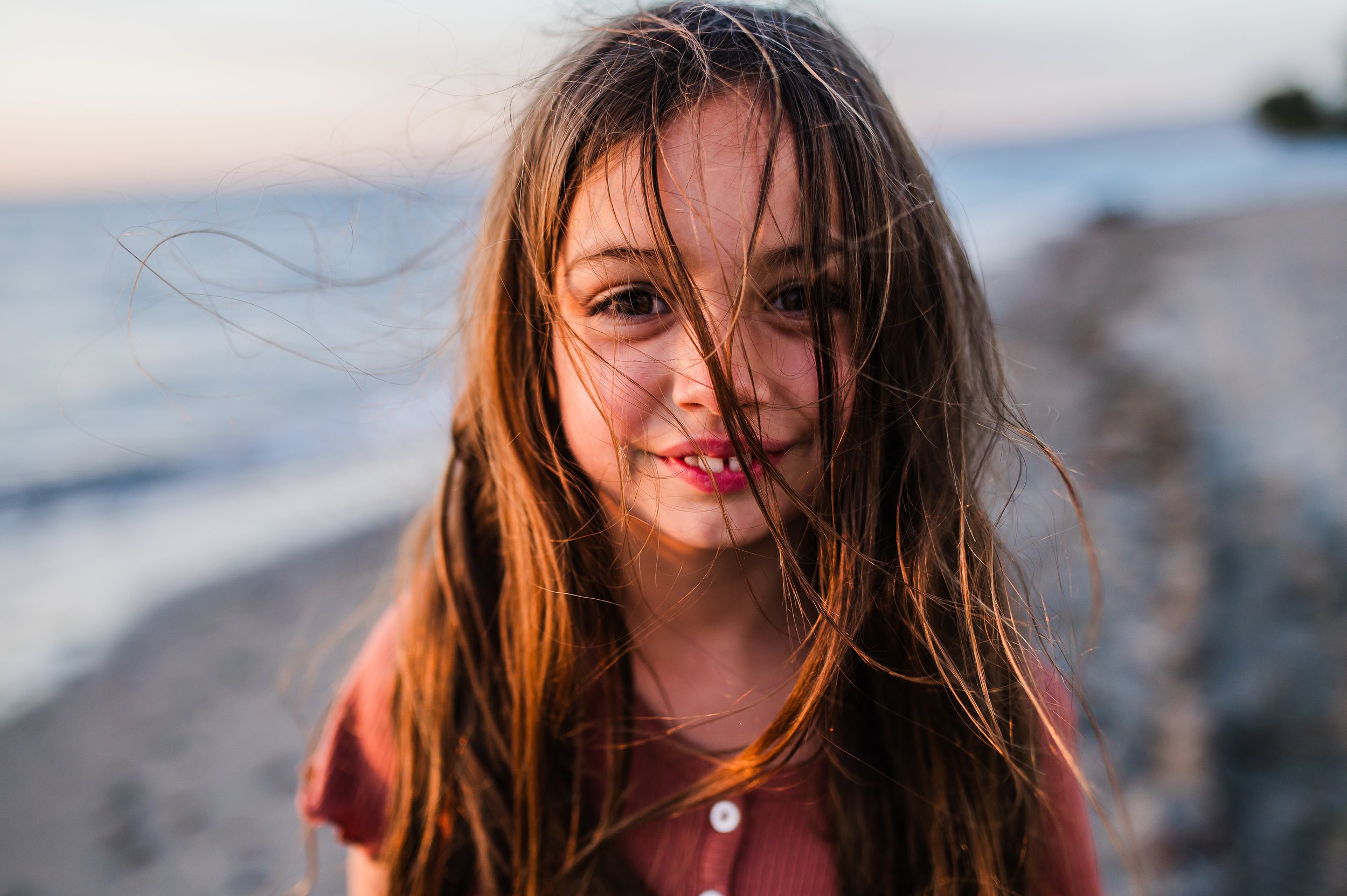 A young girl with long, wet hair smiling at the camera on a beach at sunset.