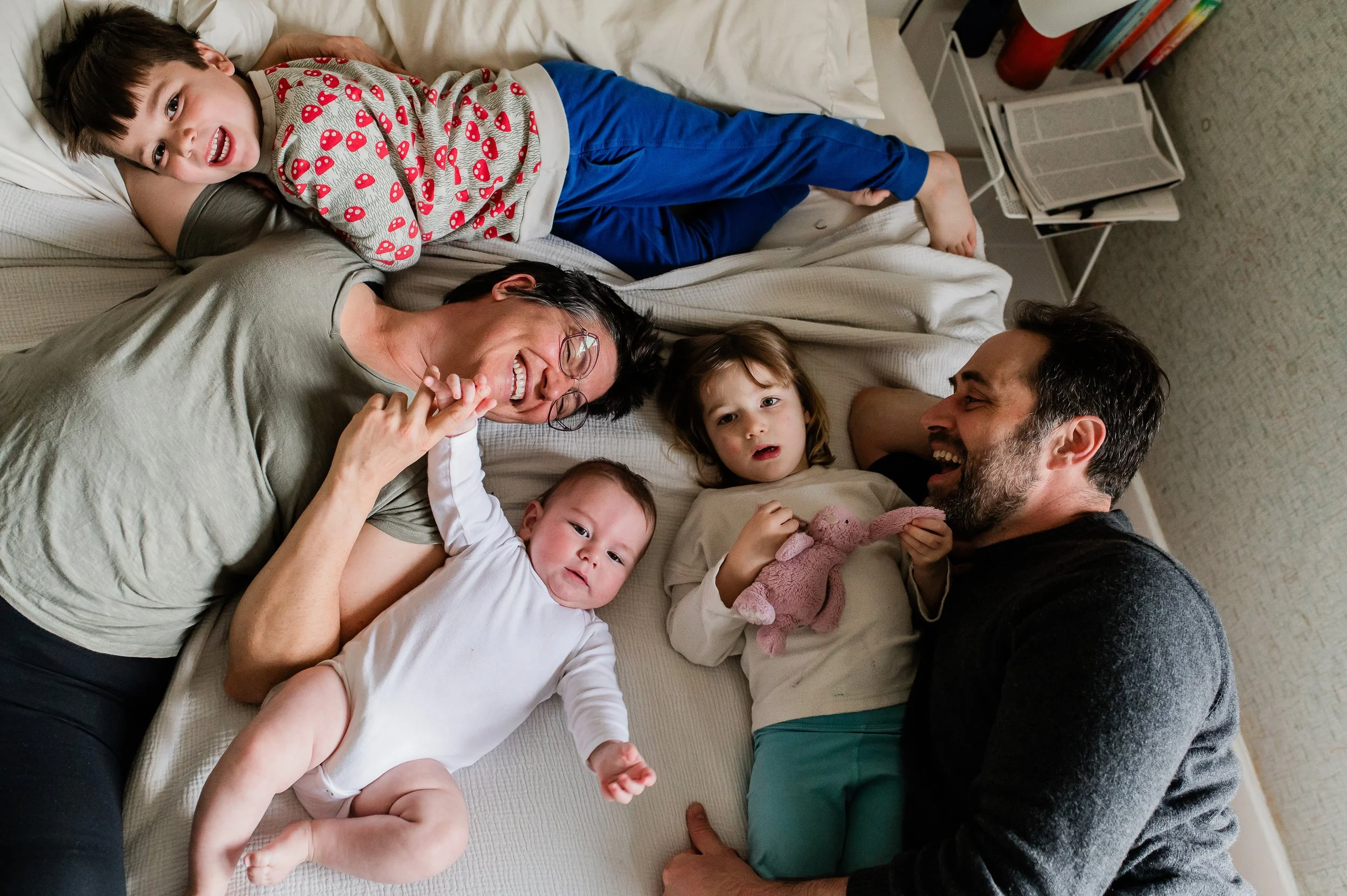 A family of six lying on a bed, engaging in playful interaction. The boy, with short dark hair, is smiling and looking at the camera while wearing a long-sleeve shirt with red mushroom patterns and blue pants. The woman, with glasses and dark hair, i