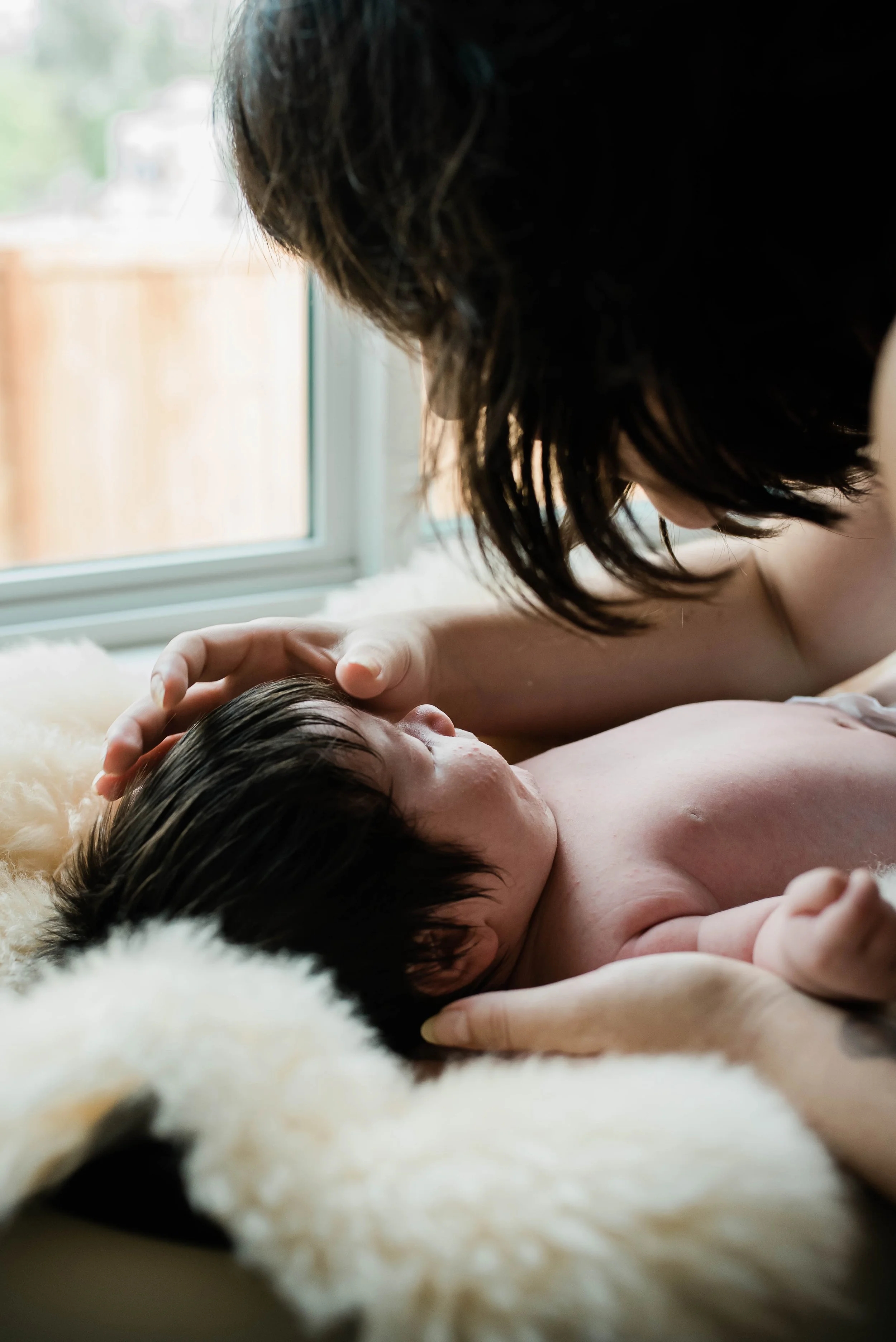 A mother and her baby lying on a soft, furry blanket near a window, with the mother gently touching the baby's head.