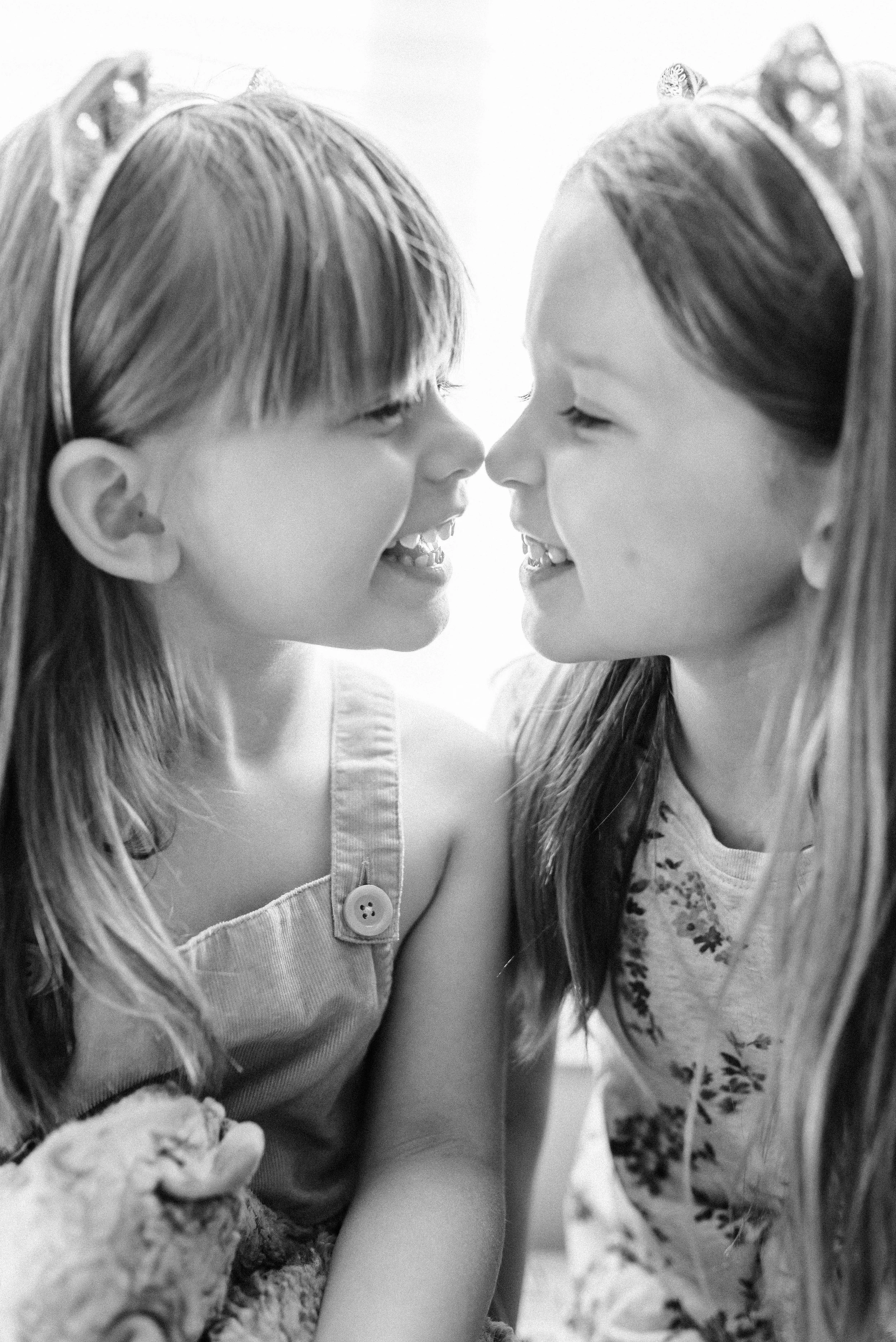 Two young girls with long hair and headbands, facing each other closely, smiling and touching noses, in a black-and-white photo.