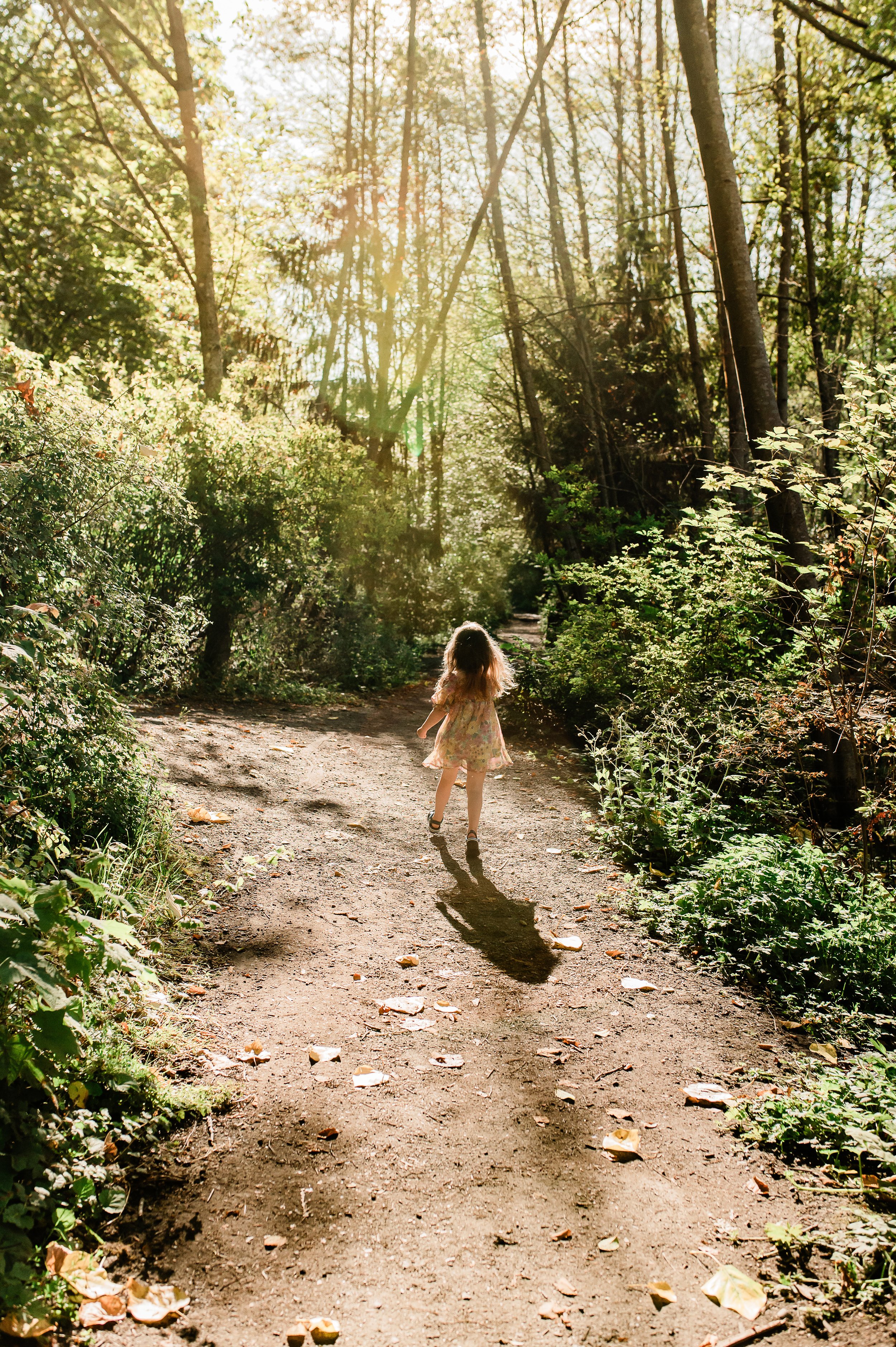 A young girl walking on a dirt trail in a wooded forest with sunlight streaming through the trees.