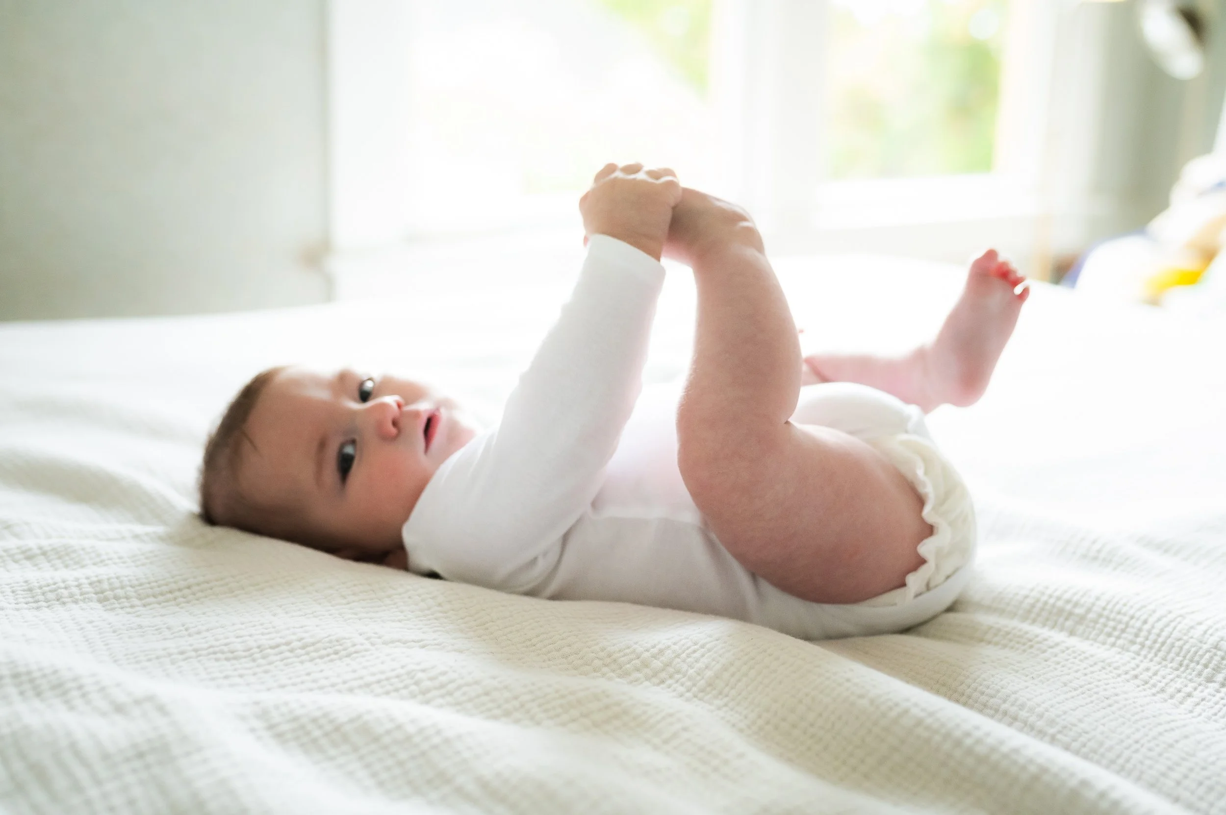 A baby lying on a bed, holding up its legs and looking at the camera with a curious expression, in a room with sunlight coming through a window.