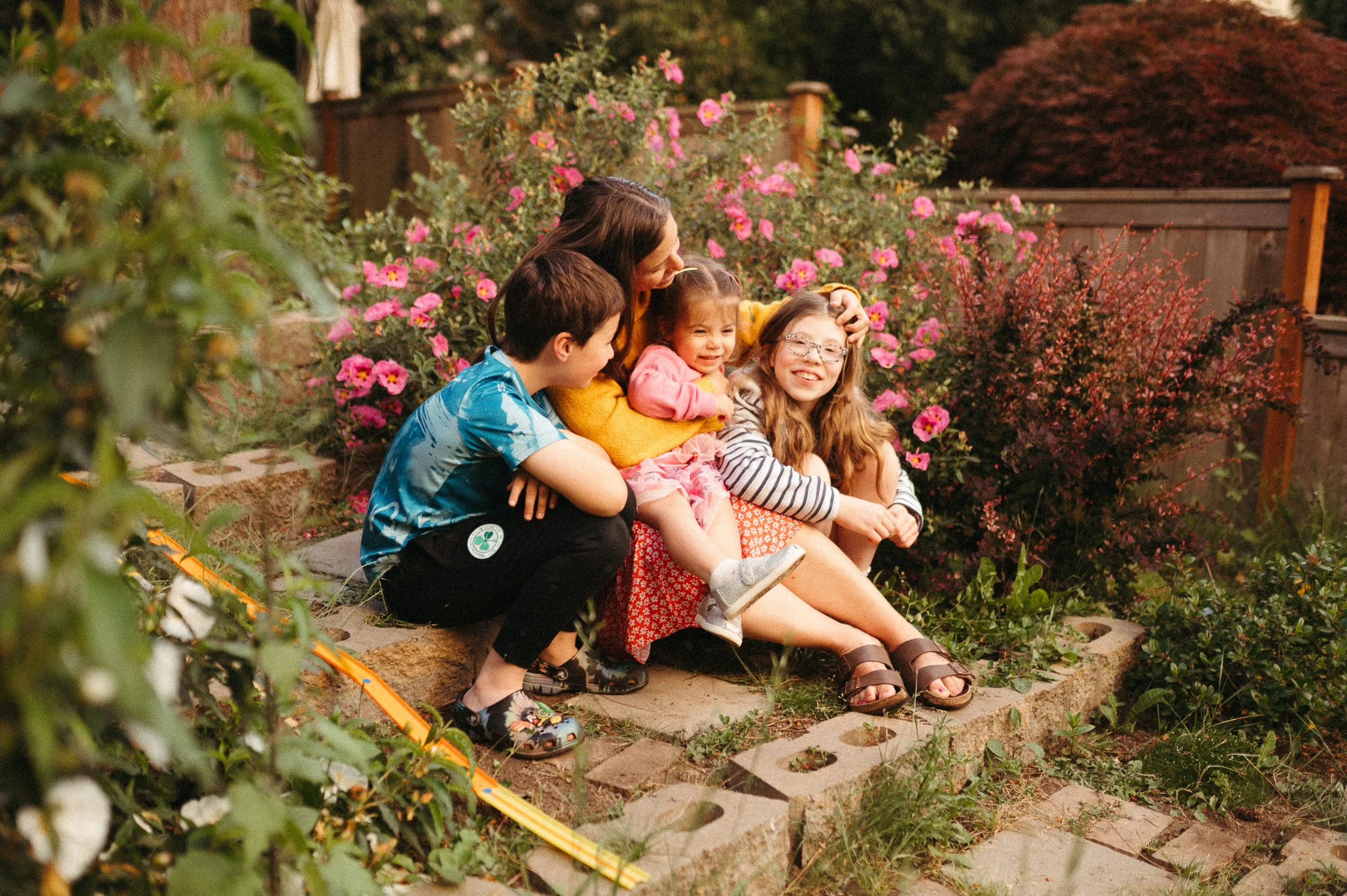 A woman and three children sitting on a brick garden ledge surrounded by pink flowers and greenery, smiling and enjoying each other's company during sunset.