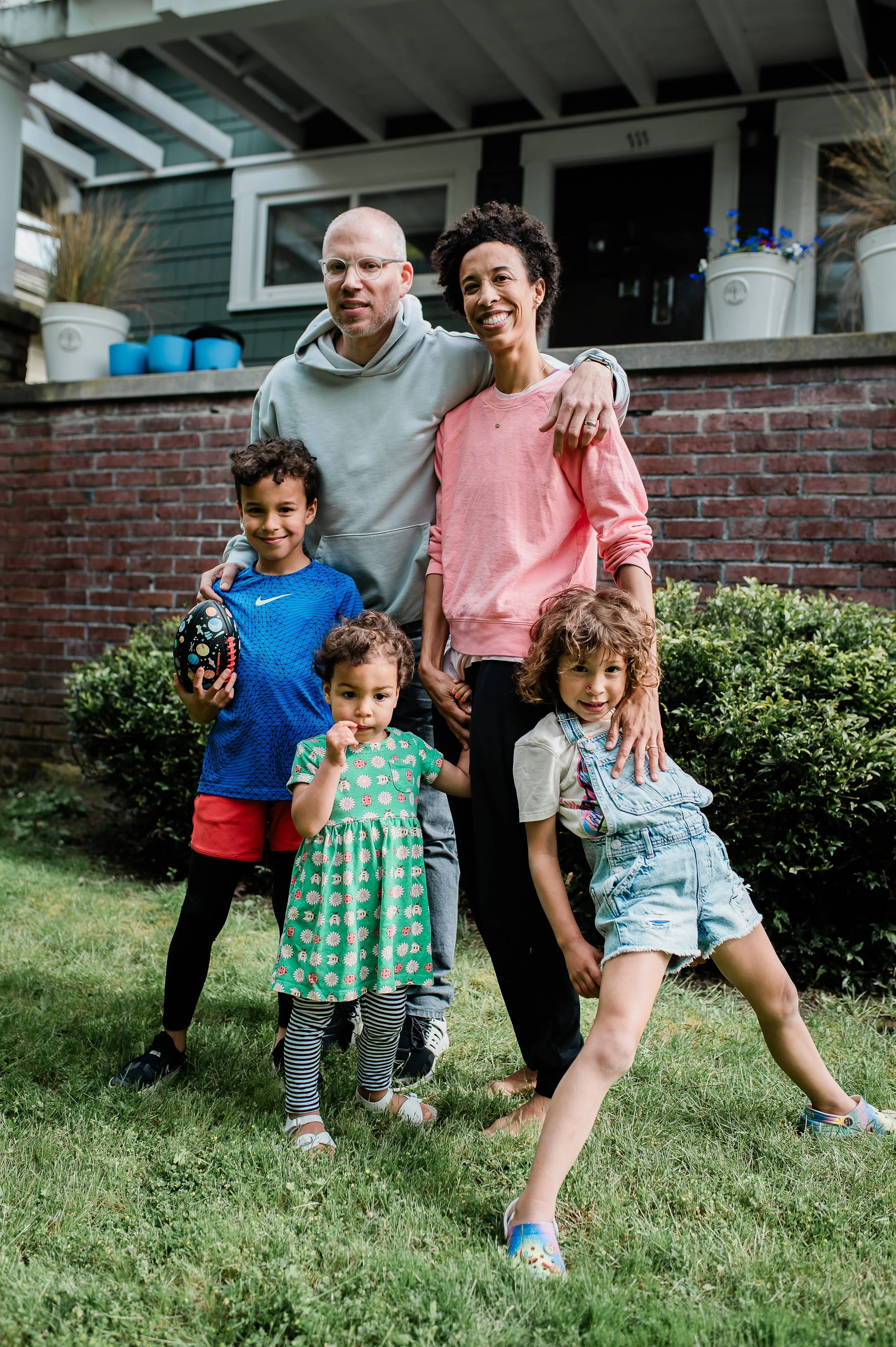 A group of six people, including three children and three adults, standing on a lawn in front of a brick wall and a house. The children are holding a football, and everyone is smiling, celebrating outdoor.