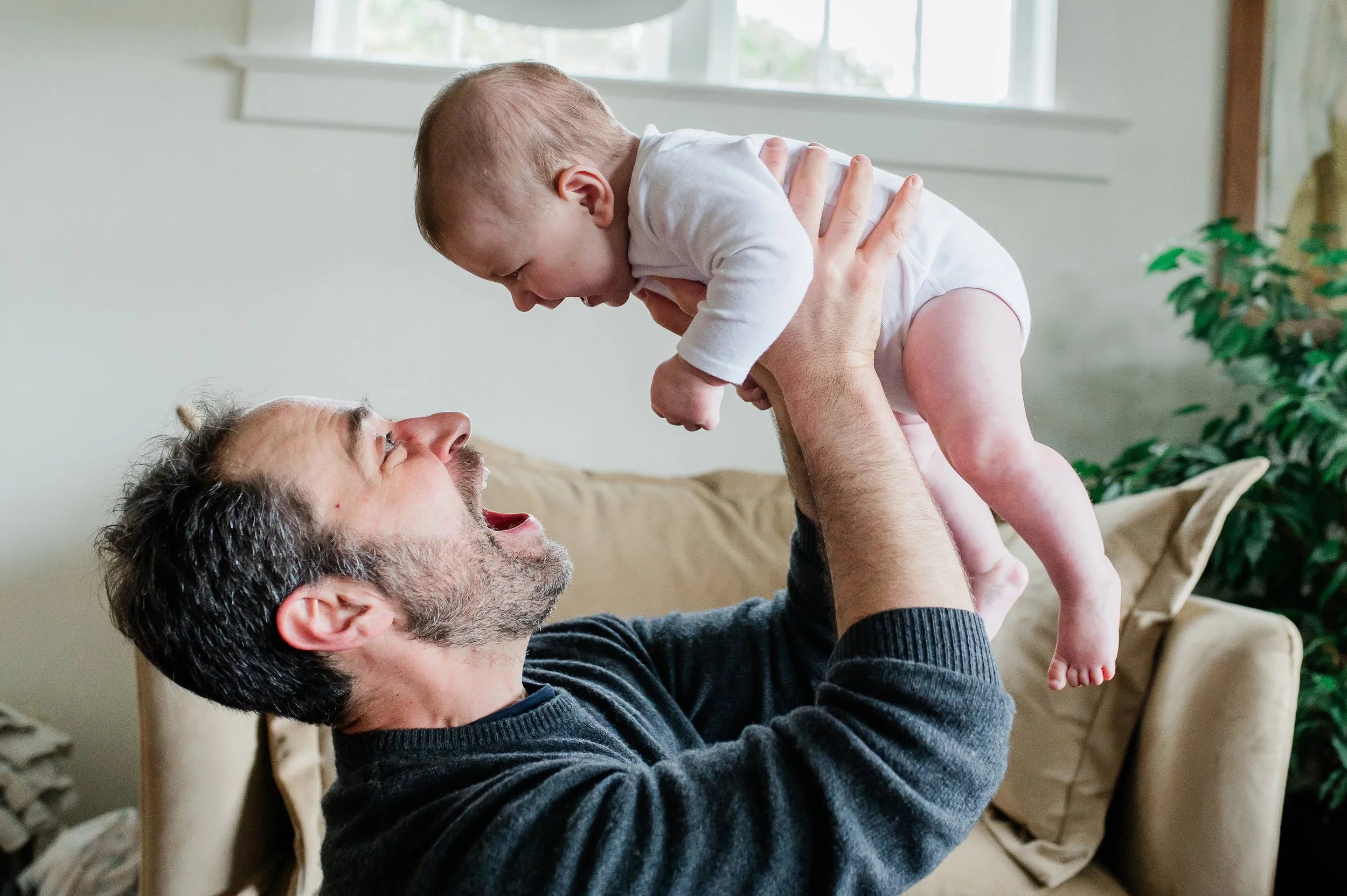 A man with gray hair and a beard lifting a baby, who is wearing a white onesie, in a living room with a window, a beige couch, and green plants.