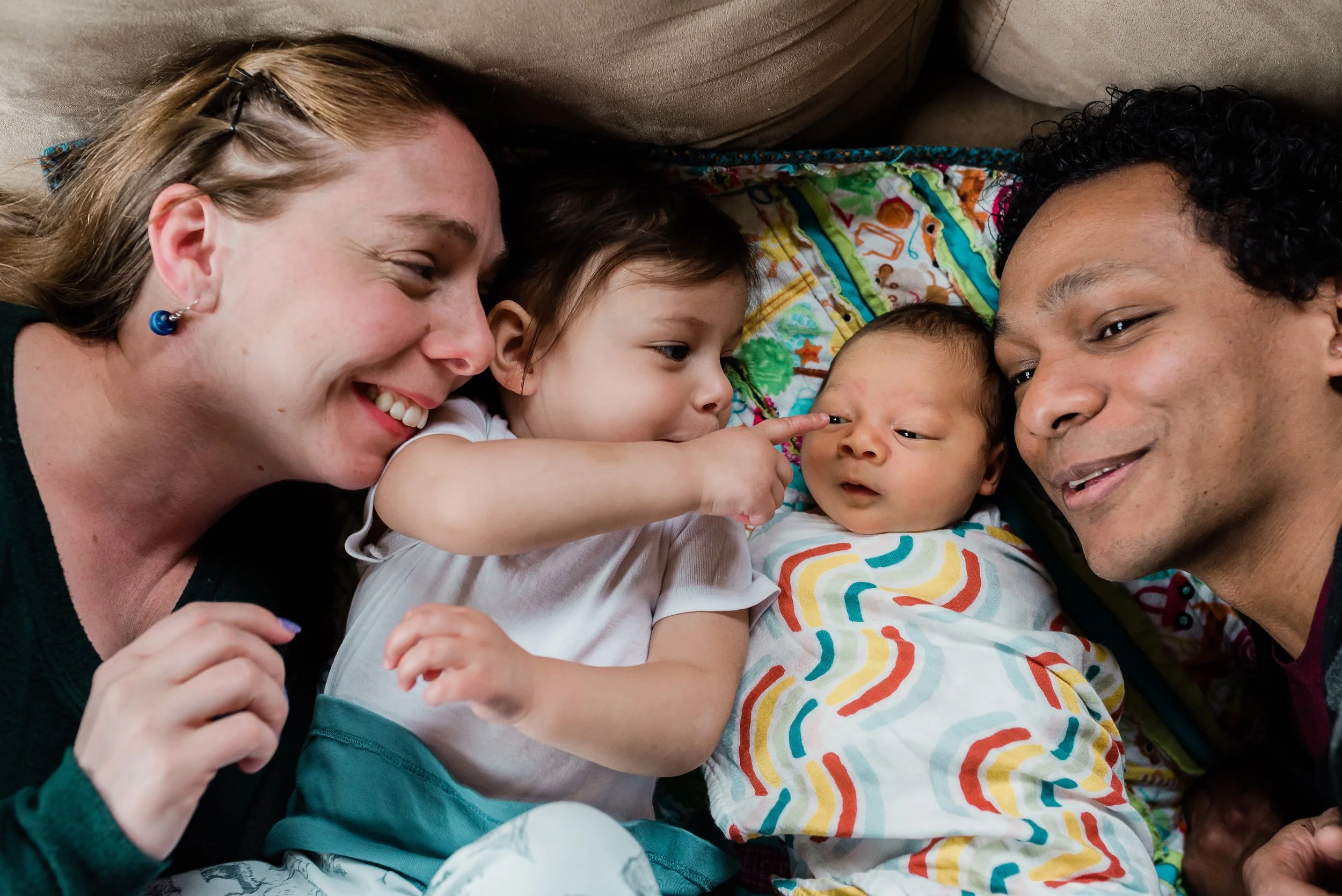 Four people lying closely together on a couch, smiling and interacting with a small baby, with a colorful blanket in the background.