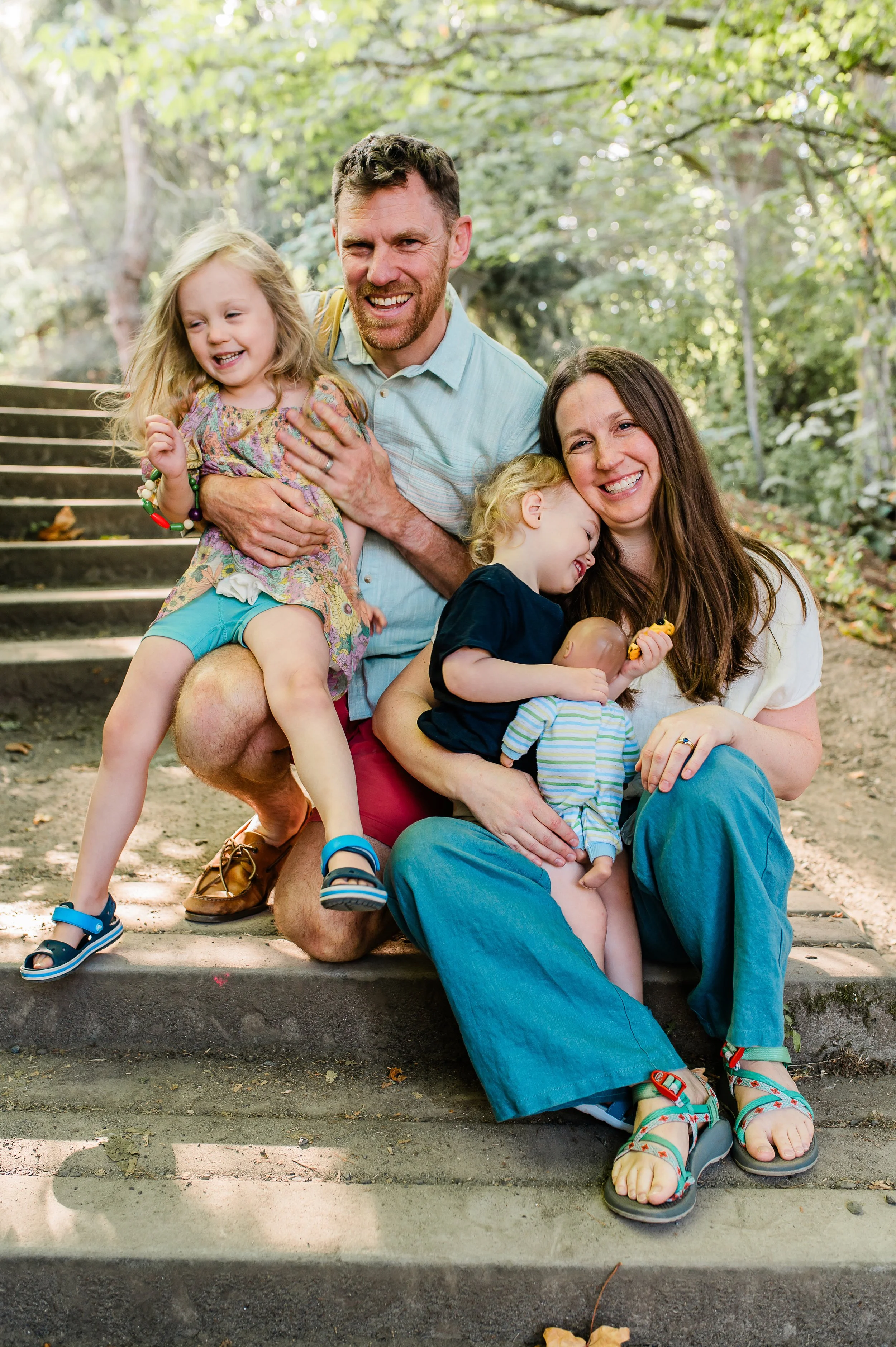 A family of four enjoying time outdoors in a wooded area, sitting on steps, smiling and laughing. The father has a beard and is wearing a light blue shirt, holding a young girl with long blonde hair who is wearing a colorful dress. The mother has long brown hair and is wearing a white shirt, holding a boy with blonde hair who is wearing a dark shirt, and is holding a small toy. They appear happy and relaxed.
