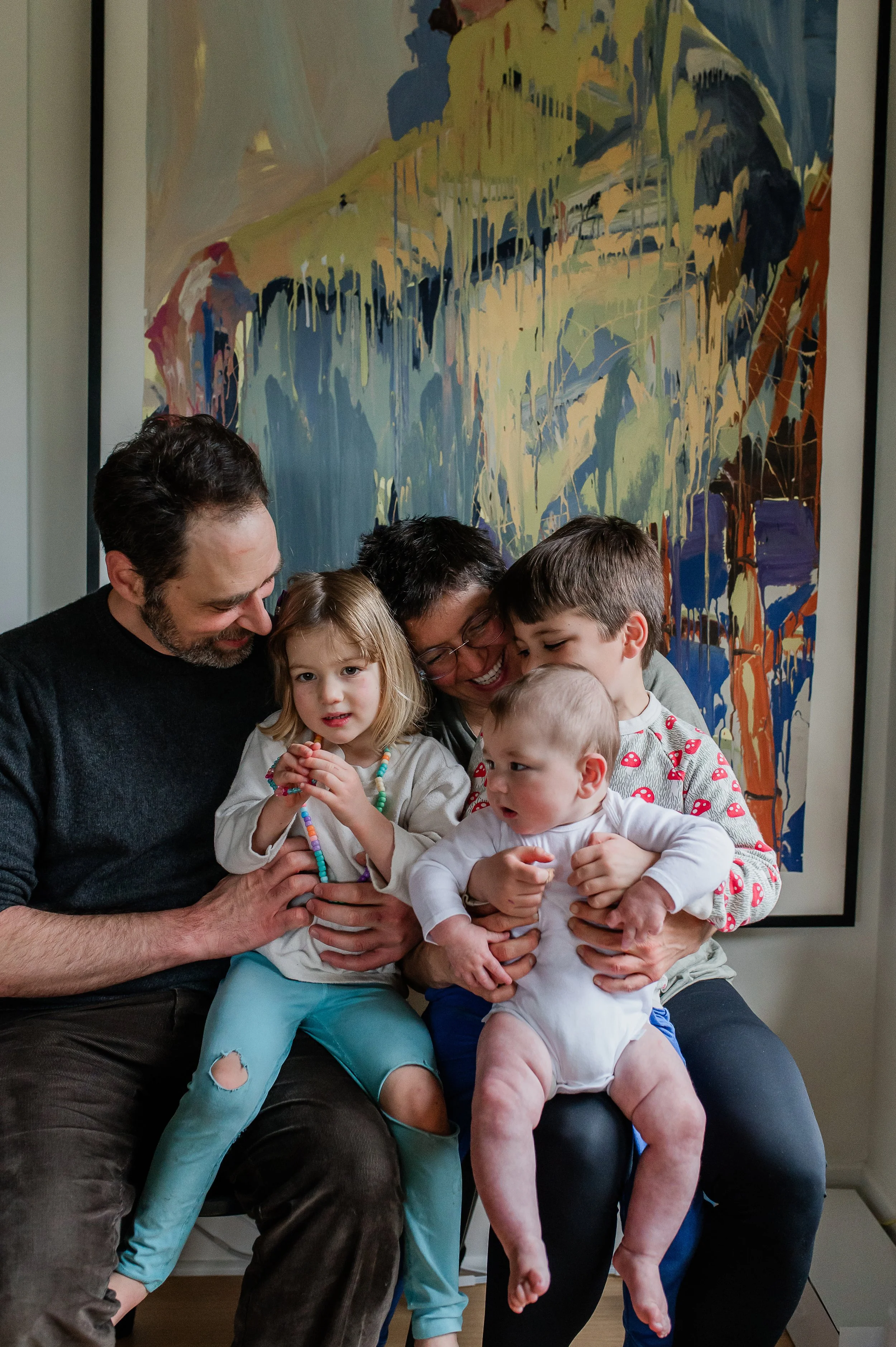 A group of six people, including two adult men, two women, and three children, gathered together, smiling and appearing happy. They are sitting closely in front of a large abstract painting with various colors in a room.