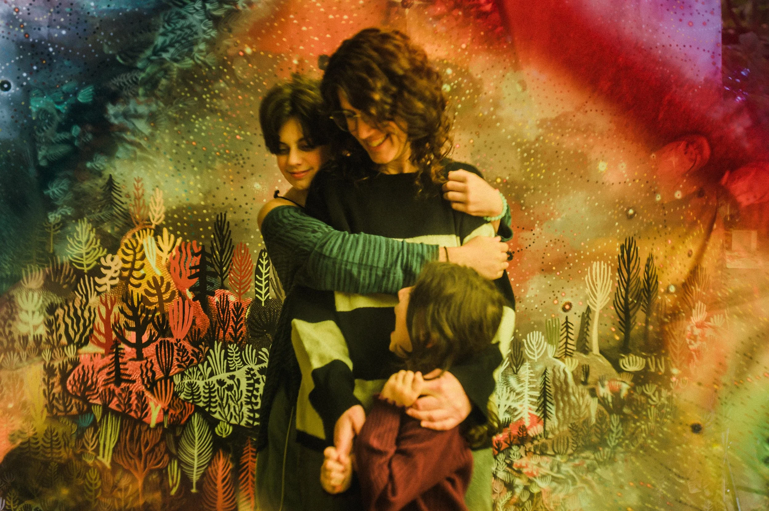 A woman with curly hair and glasses hugs a young girl in a burgundy sweater, while another child in a striped shirt hugs a person with their back to the camera. They are in front of a colorful, abstract background with patterns of trees and small dot