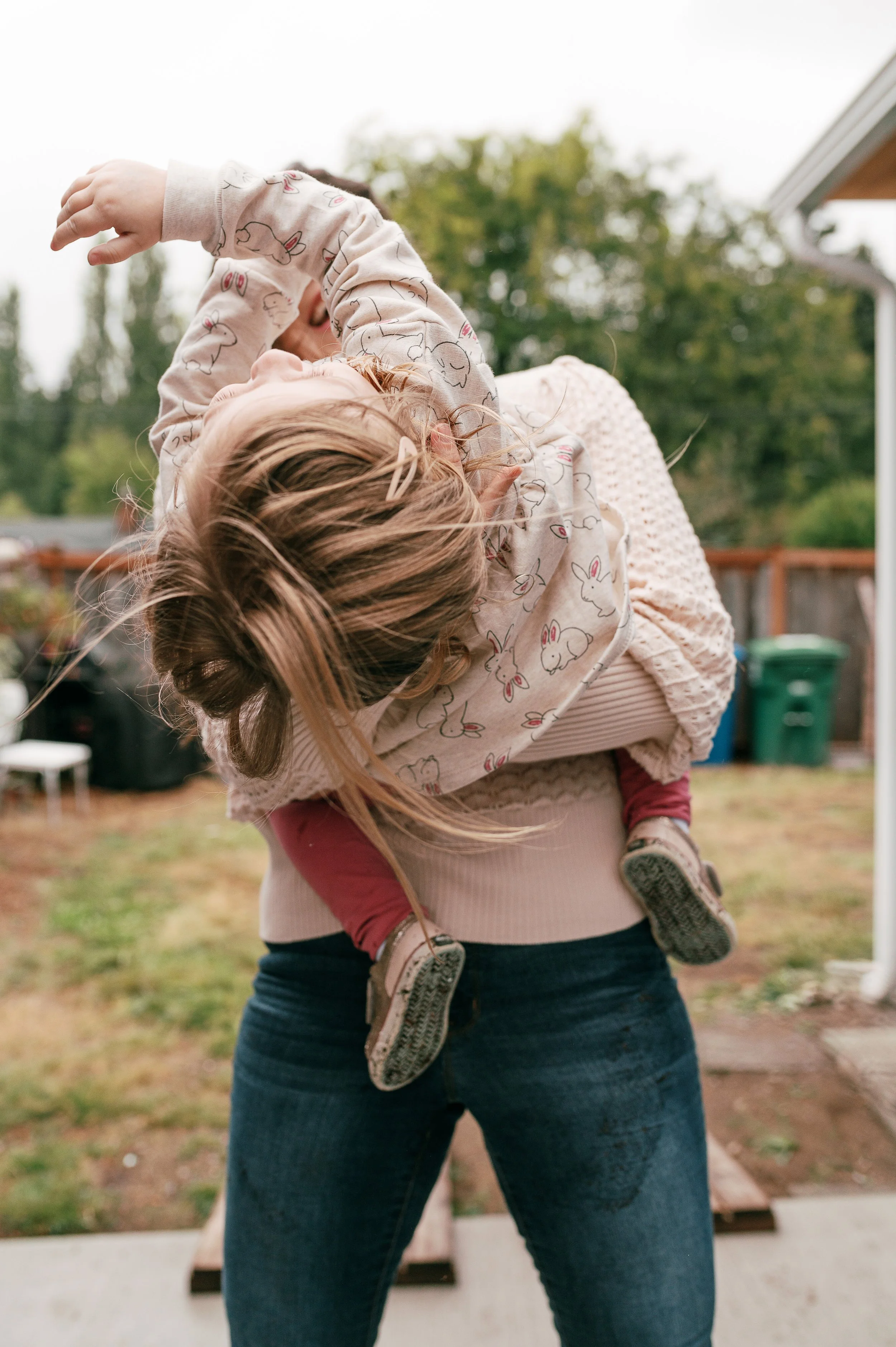 A woman holding a young girl outdoors, with trees and a house in the background, on a cloudy day.