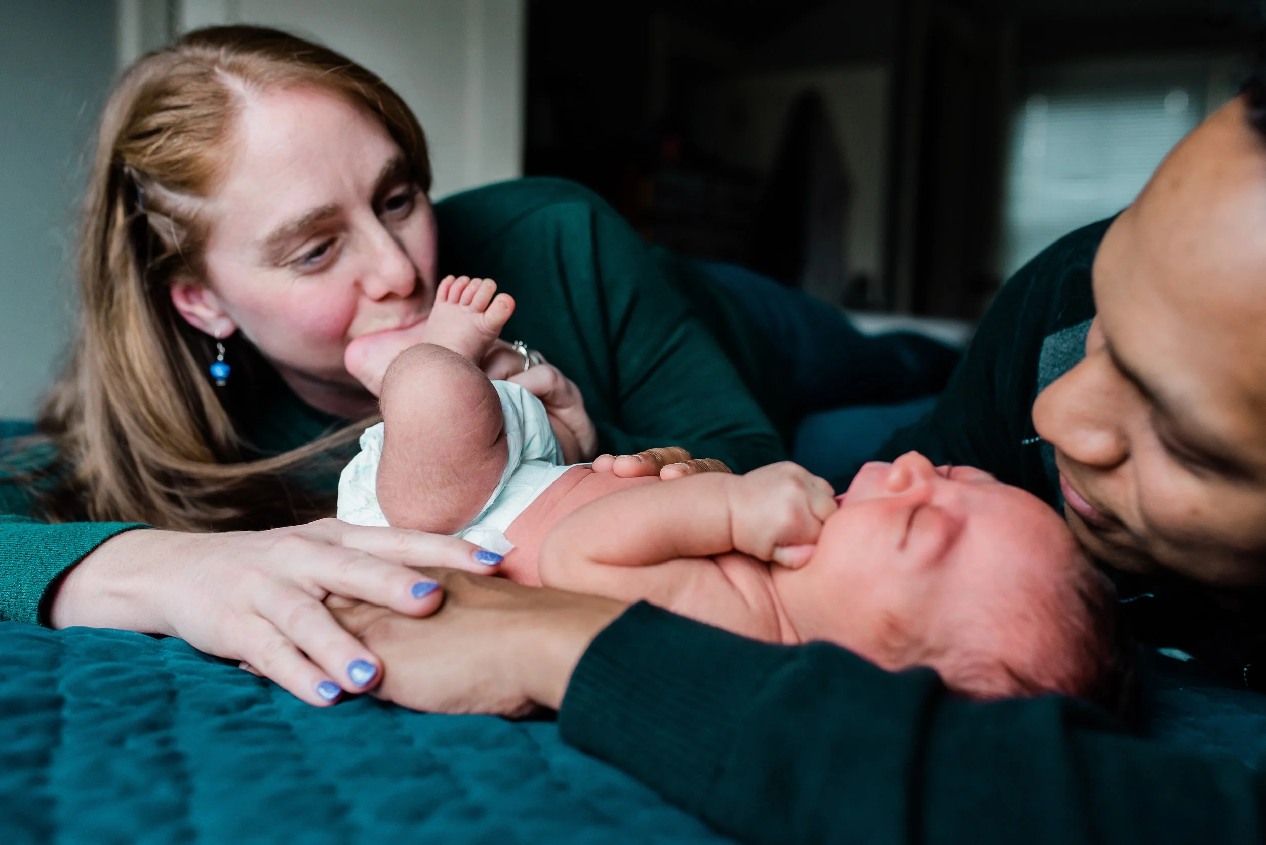 A woman with long reddish-brown hair and earrings is lying on her side, gently holding a newborn baby on a dark blue bedspread. The woman is smiling and looking at the baby, who is touching her face with their hand. The scene appears intimate and ten