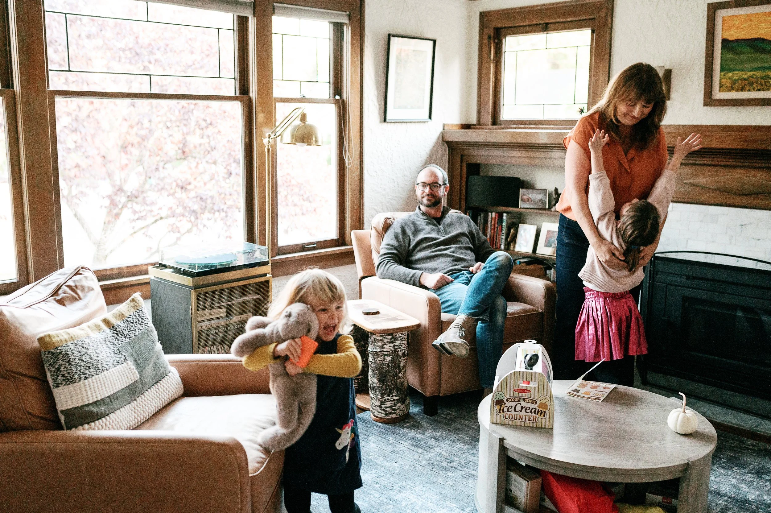 a family dances in their living room.   A child reaches for her mother and a toddler hugs her stuffed animal.