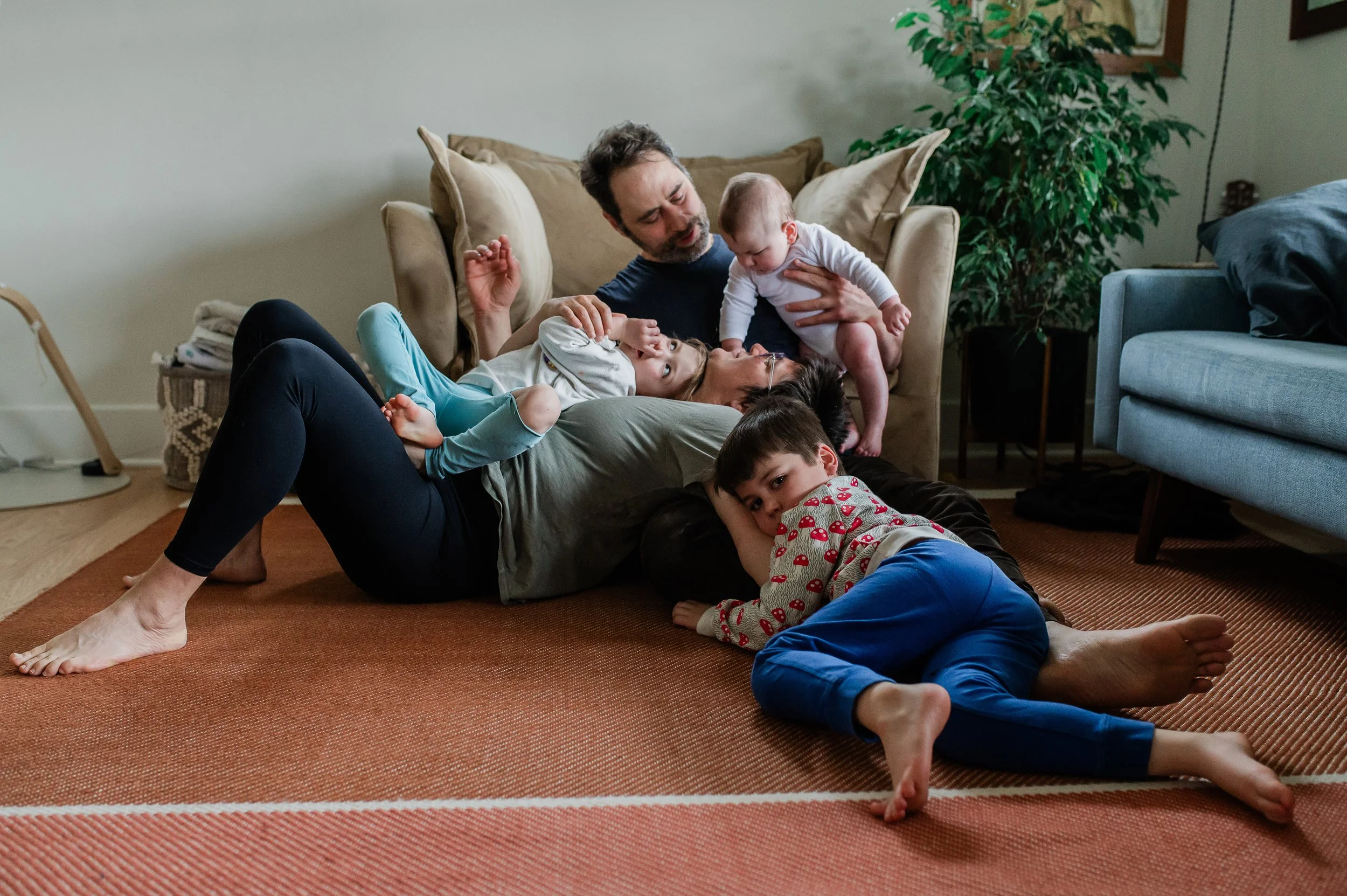 A family of four lying on a living room rug, playing and relaxing together. The father and children are engaging in a playful moment on the floor, with a sofa, a large plant, and wall decor visible in the background.
