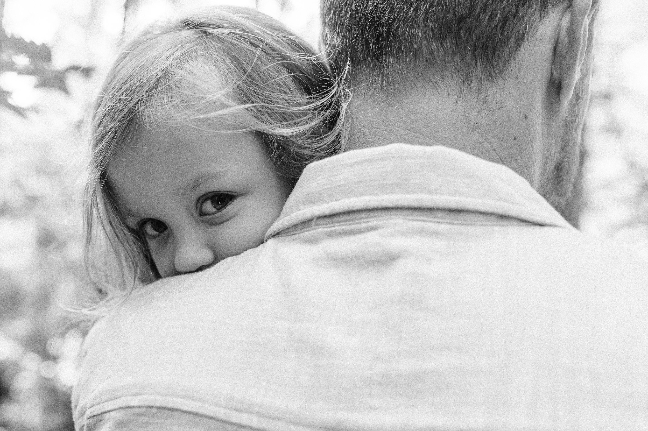 Close-up black and white photo of a child resting their head on an adult's shoulder, with their face partially visible and eye looking towards the camera.