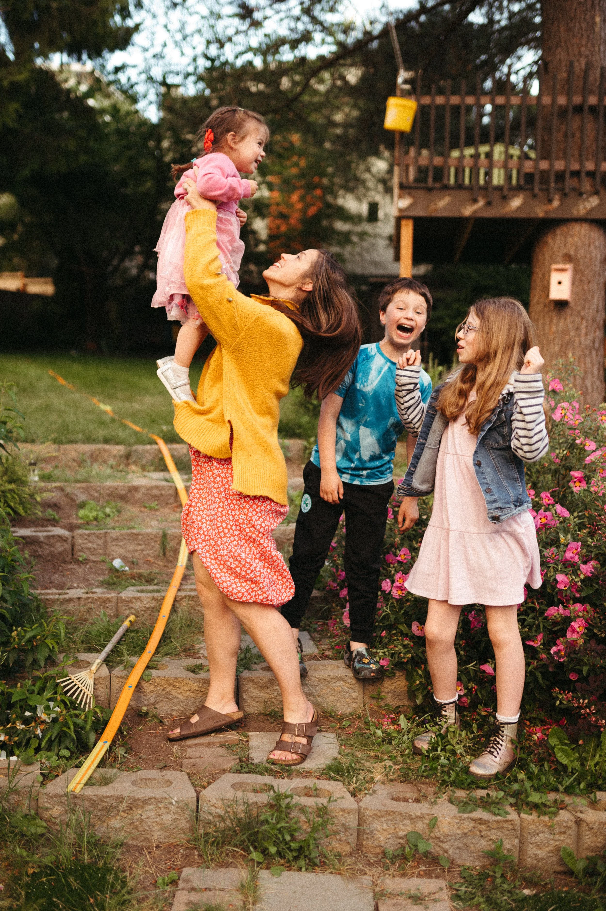 A woman wearing a yellow sweater and patterned skirt lifts a young girl in a pink dress with a red hair bow, smiling at each other. Two other children, a boy and a girl, stand nearby, laughing and talking. They are outdoors in a backyard with a treehouse in the background, surrounded by flowers and greenery.