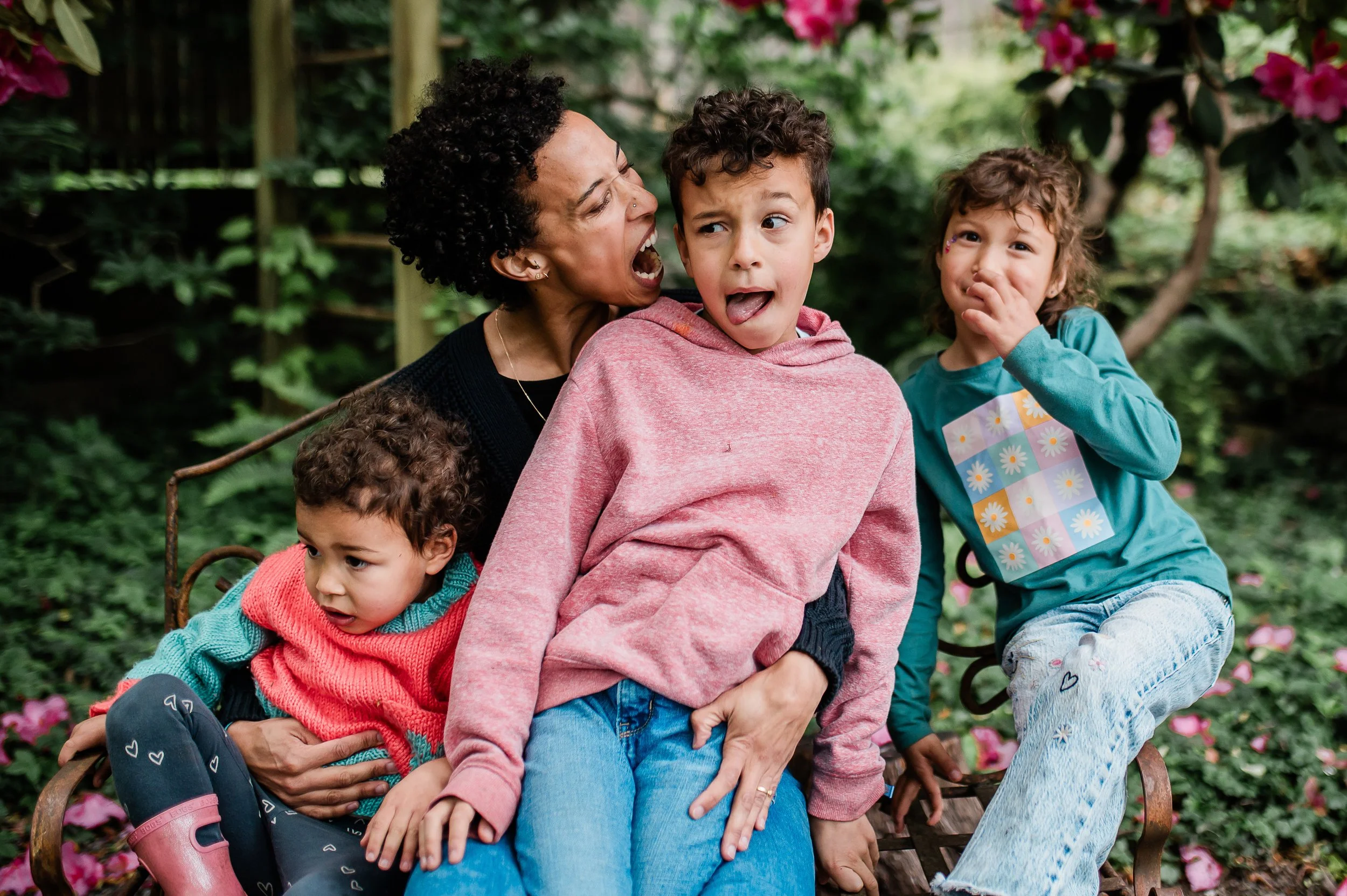 A woman with short curly black hair and a nose piercing sits on a wooden bench outdoors with four children surrounded by pink flowers and green foliage.