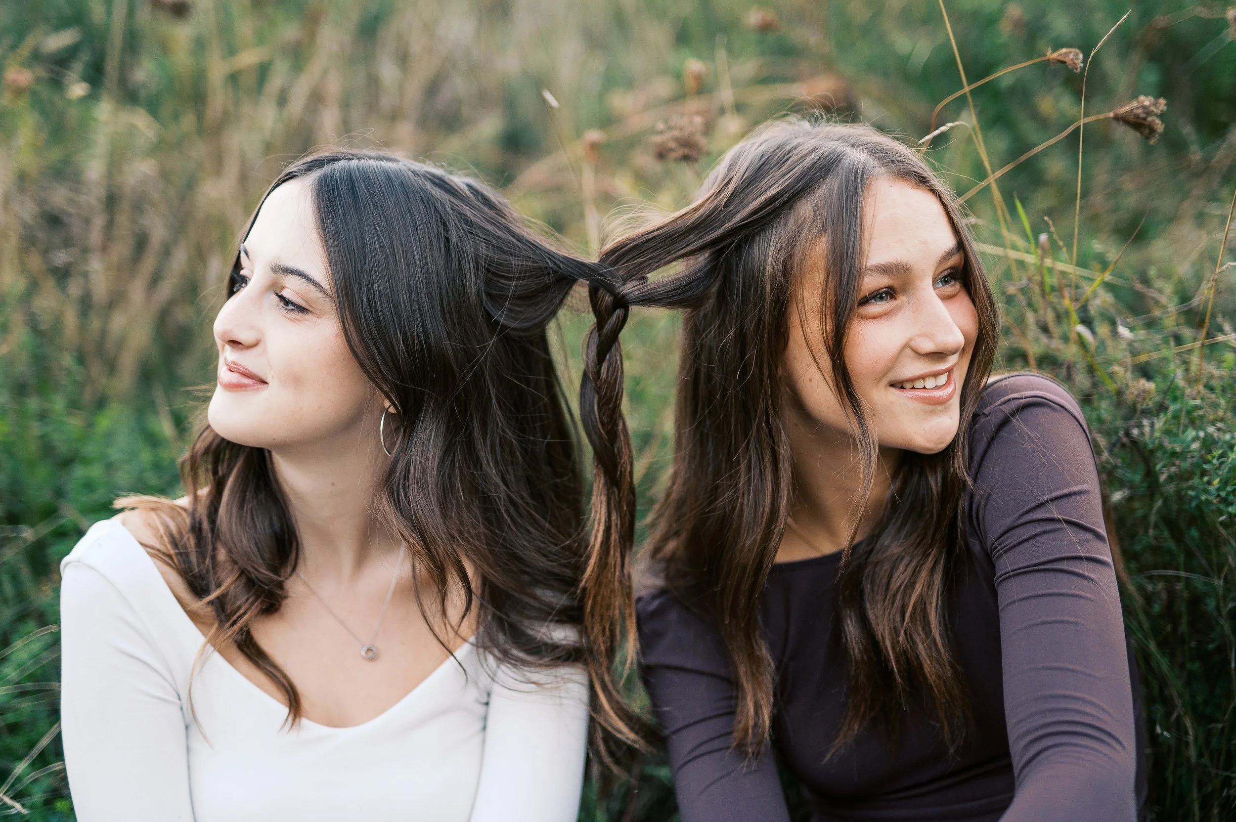 Two young women sit back to back in a natural setting with tall grasses, with their hair intertwined.