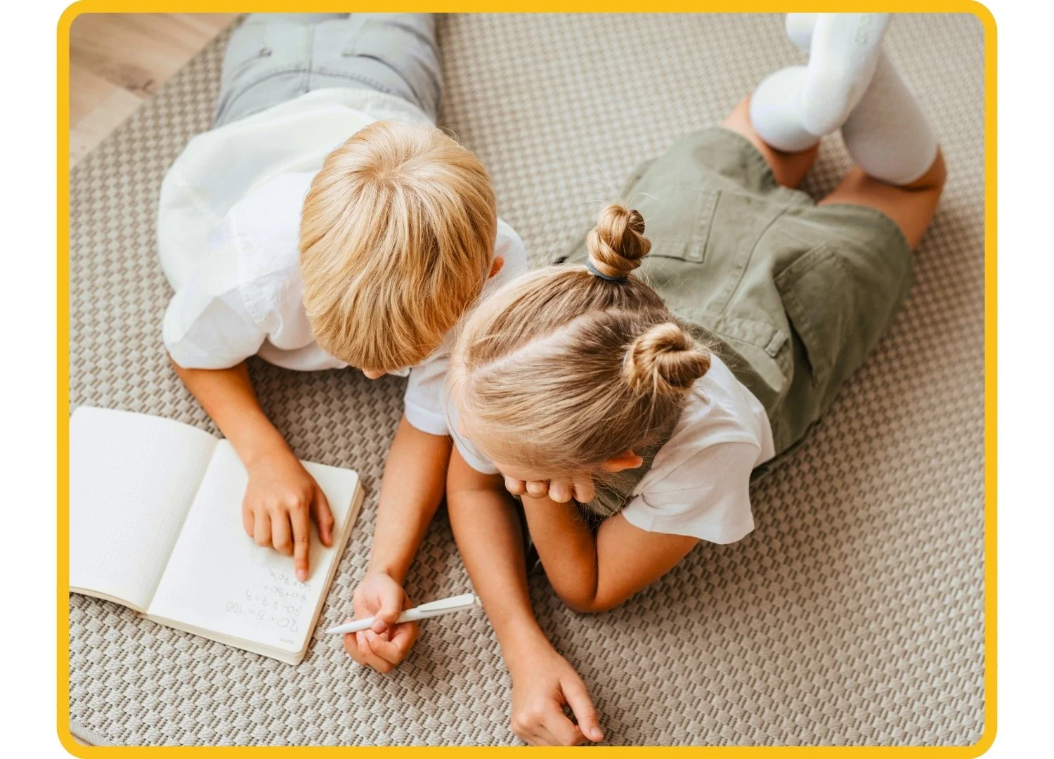Two children lying on the floor, one is writing in a notebook