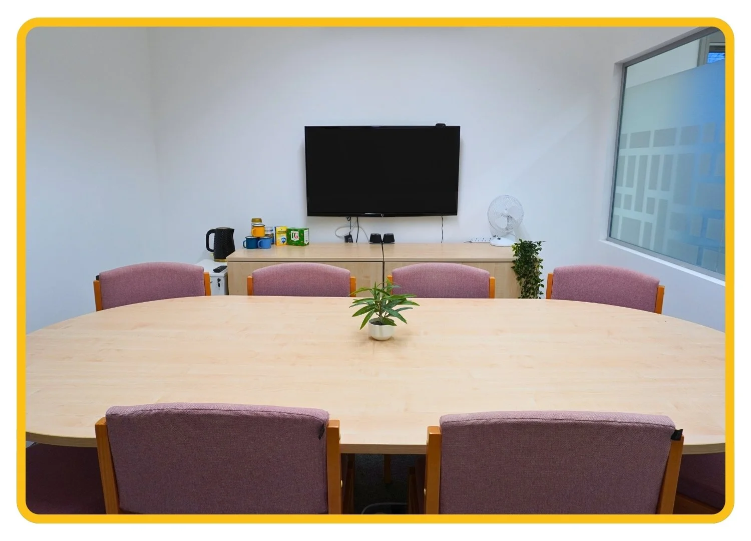 Board room at Cambridge (Stow-cum-Quy) clinic. White room with frosted window with a large central table is surrounded by pale pink chairs. A TV is mounted on the wall. Hot drinks facilities are on a cabinet.