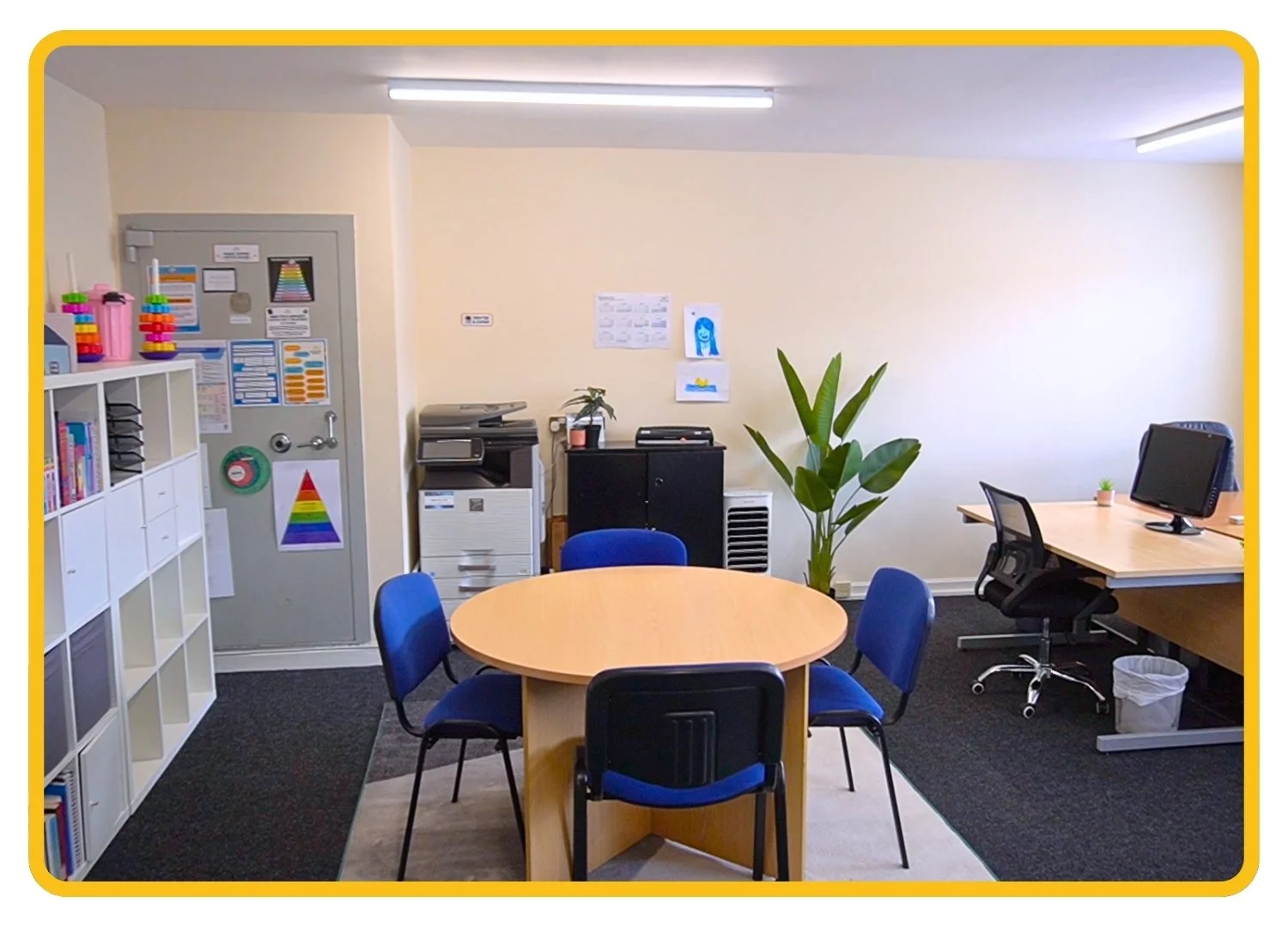 Office and co-working space at Birmingham clinic. A circular table in the middle with four chairs, a desk with laptop, large plant, white cabinets and photocopier/printer.