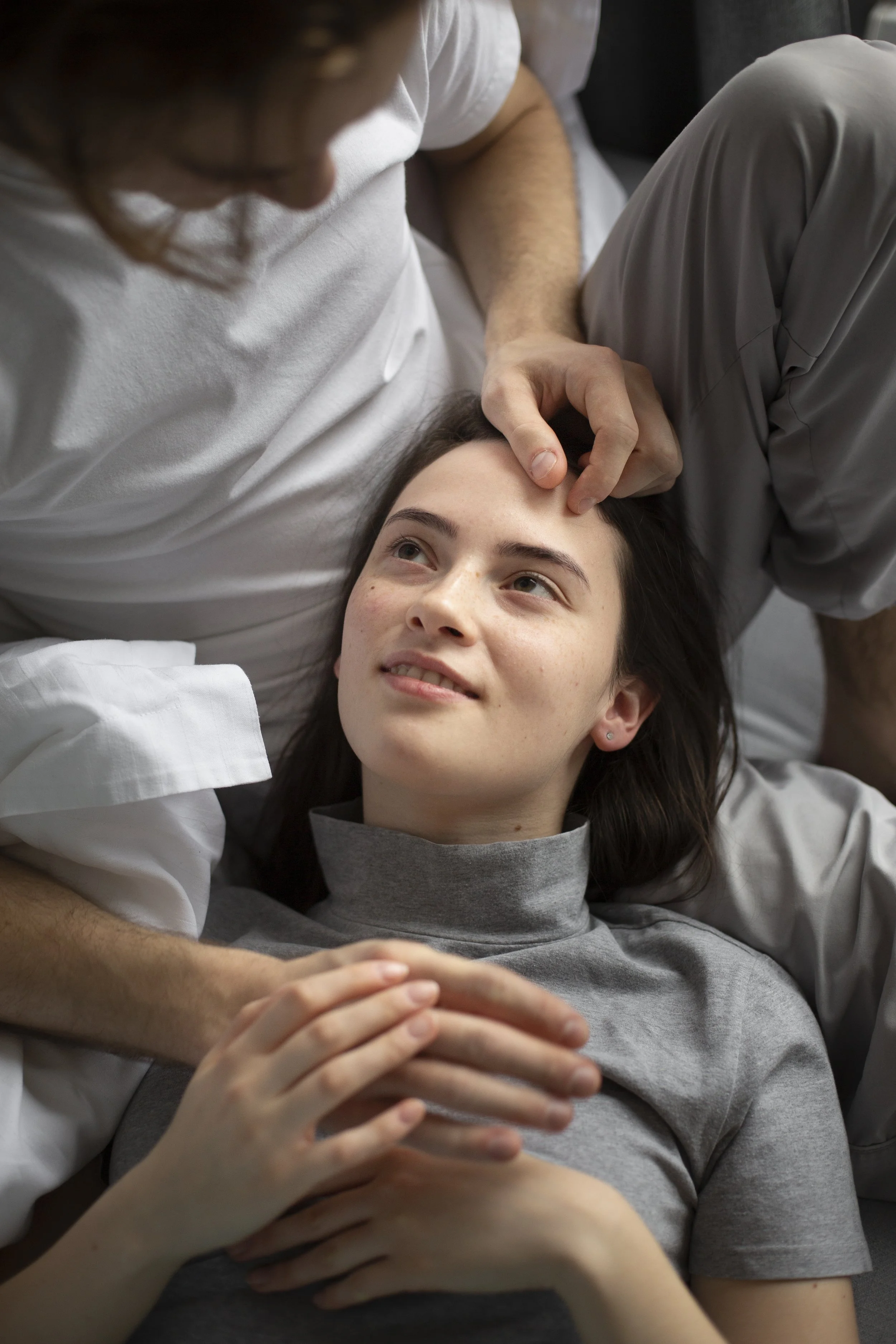 A woman lying down with her head turned to the side, looking up at a man who is gently touching her forehead and holding her hand. The man is wearing a white shirt, and the woman is wearing a gray t-shirt.