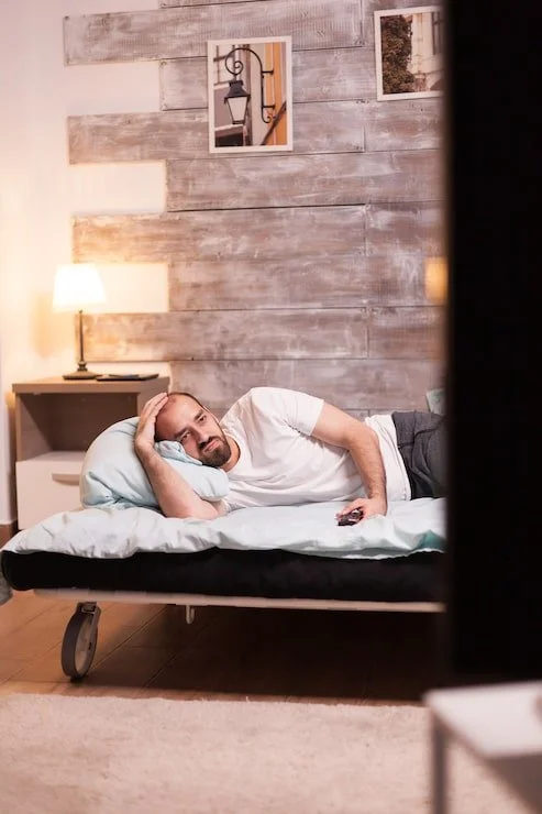 A man lying in bed with a distracted or distracted expression, resting his head on a pillow while watching television, in a cozy bedroom with wooden wall decor and lamps.